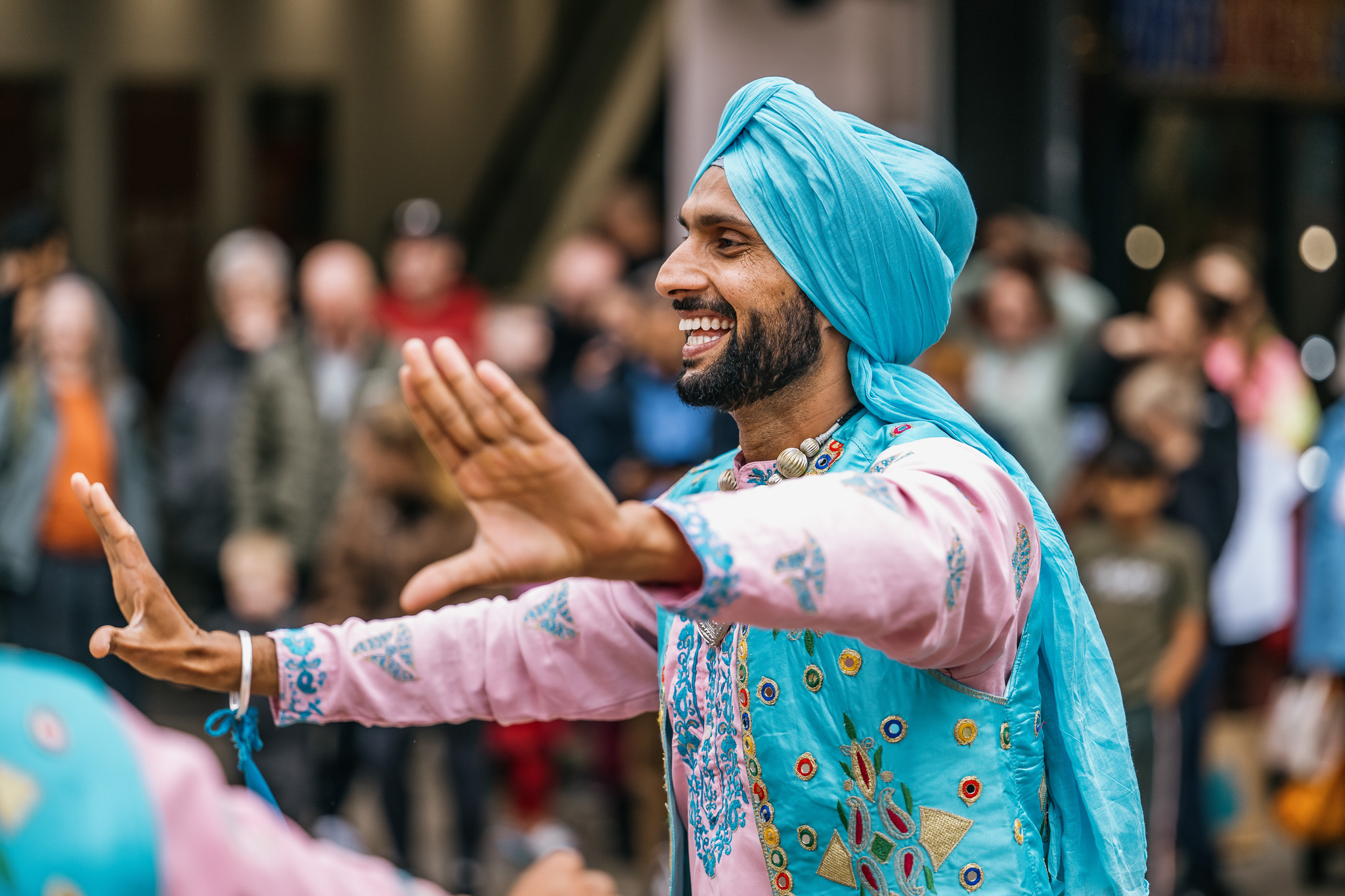 A man wearing a bright blue turban and embroidered outfit smiles and dances with his arms outstretched, performing at an outdoor event with a blurred crowd in the background.