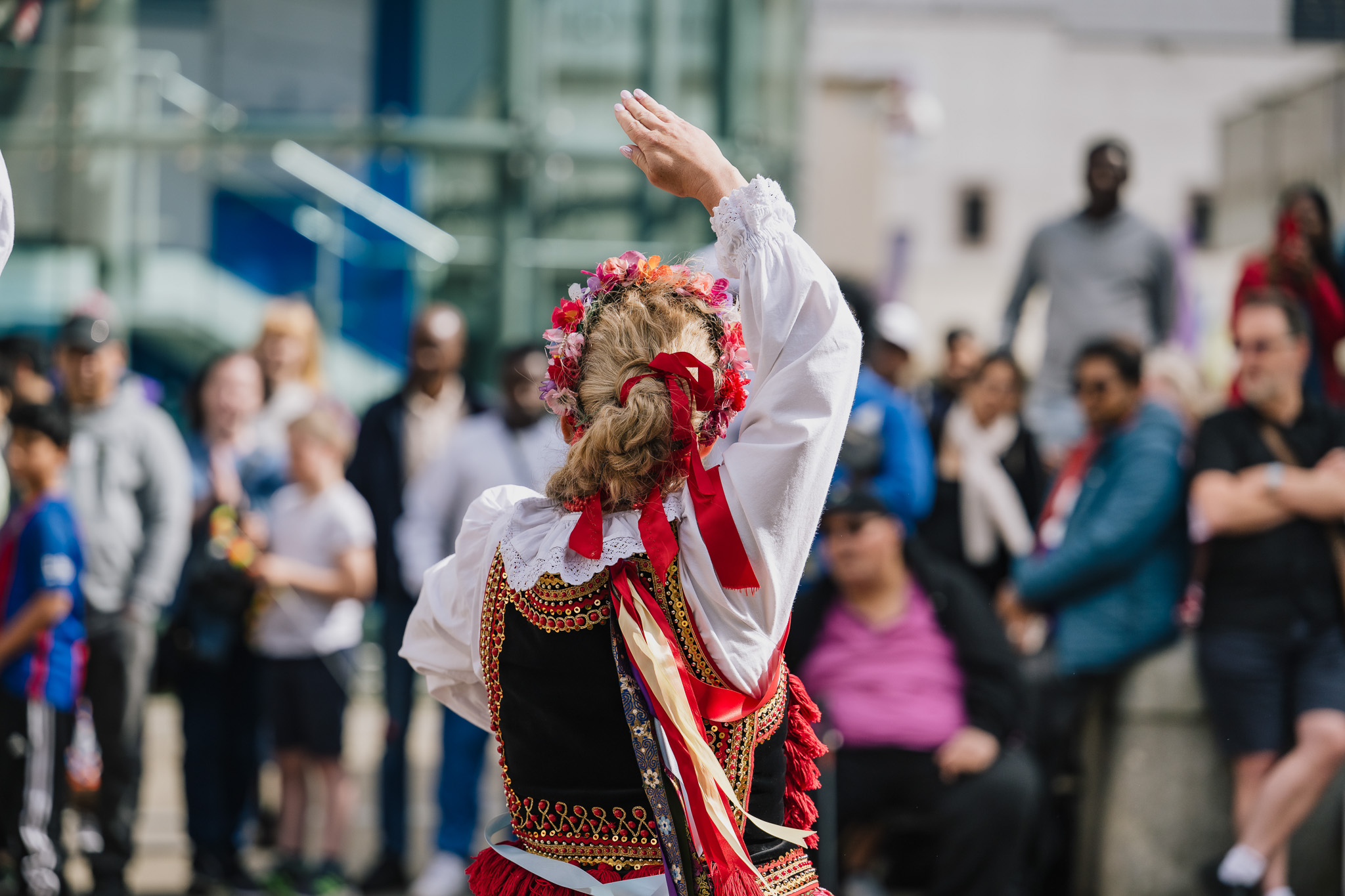 A woman in traditional folk attire with red ribbons and a floral headband dances outdoors, facing a crowd of onlookers who are watching her performance. The scene appears festive and lively.