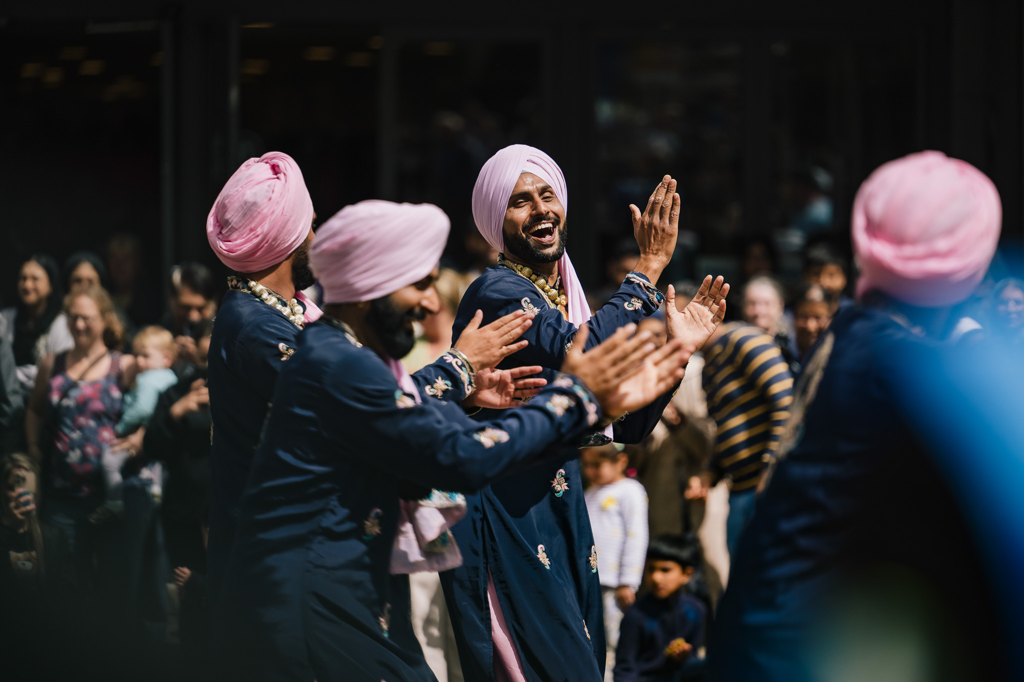 Three men in navy blue outfits and pink turbans joyfully clap and dance in front of a crowd at an outdoor event, with onlookers watching and smiling in the background.