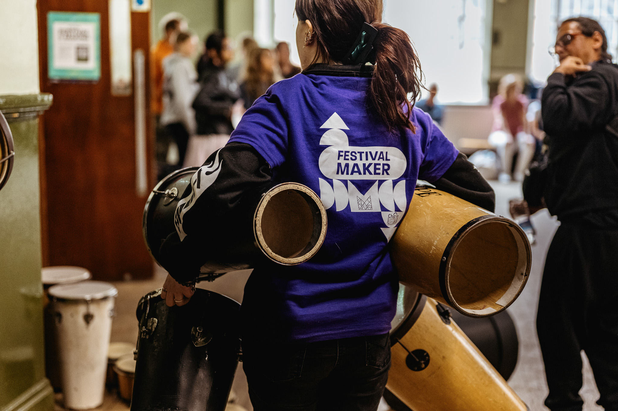 A woman wearing a blue Festival Maker shirt carries several drums through a busy indoor event space, with other people and more drums visible in the background.