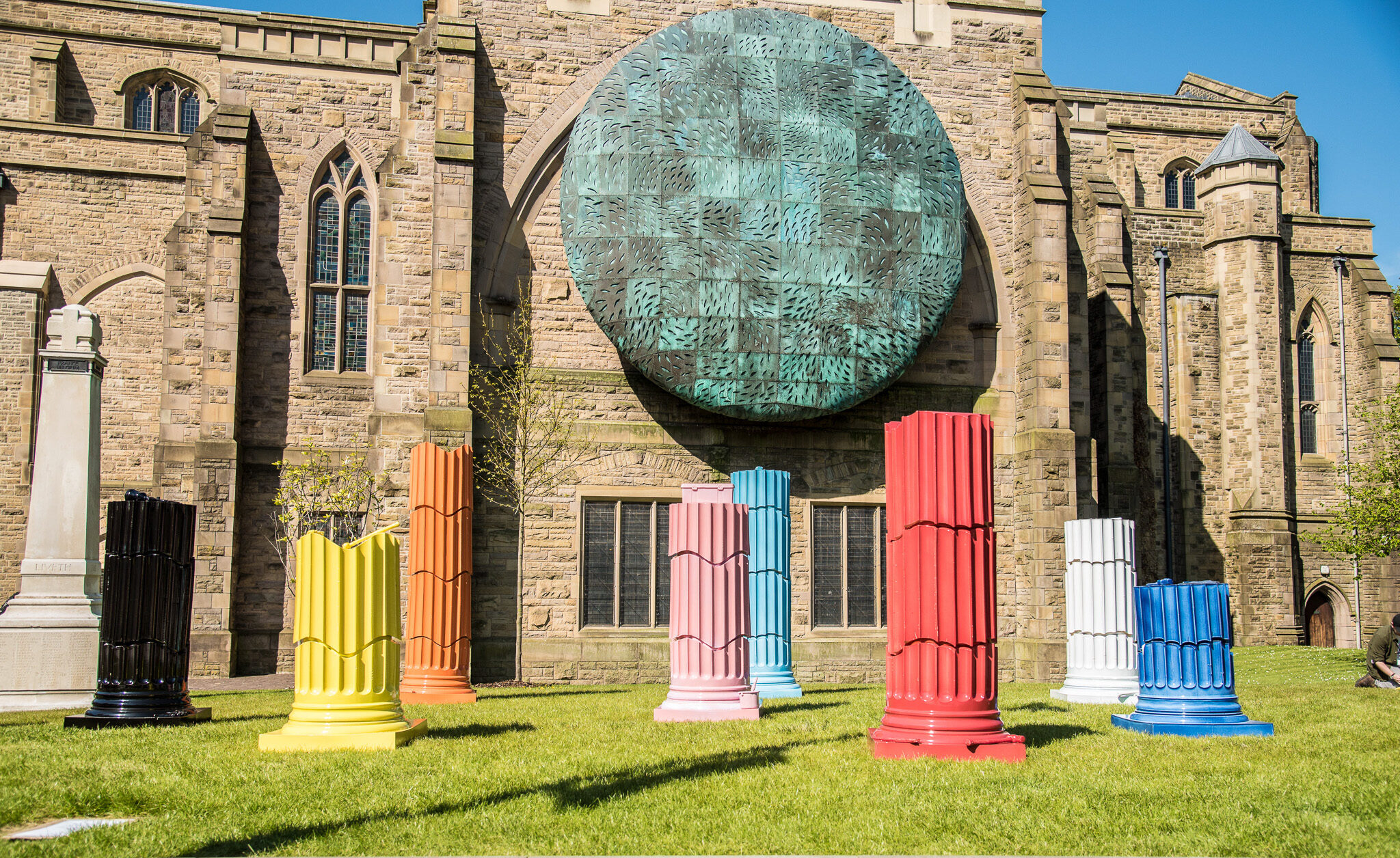 Colourful, painted column sculptures stand on grass in front of a large old stone building with arched windows and a circular green textured feature on its facade, under a clear blue sky.