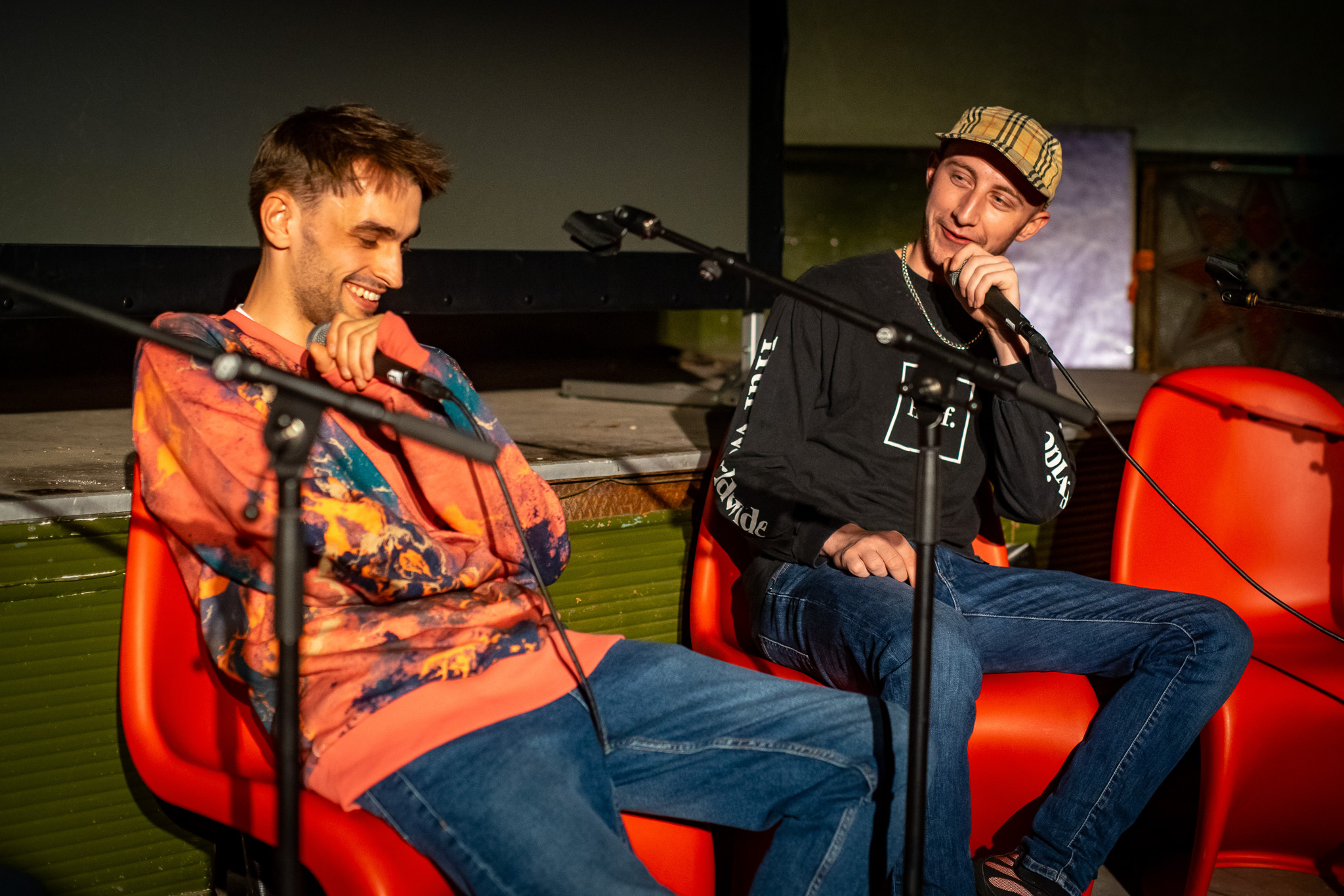 Two men sit on red chairs, smiling and speaking into microphones during what appears to be a panel discussion or interview on stage. One wears a colourful hoodie, the other a black shirt and a checked cap.