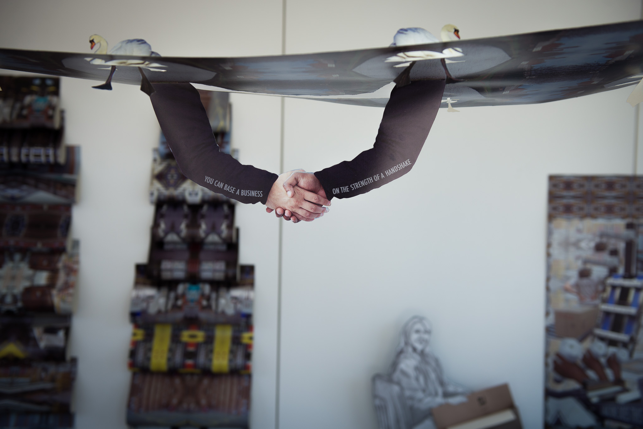Two arms extend downwards from a ceiling decorated with swan figurines, meeting in a handshake. The arms appear to emerge from holes in the ceiling. Art materials and a statue are visible in the background.