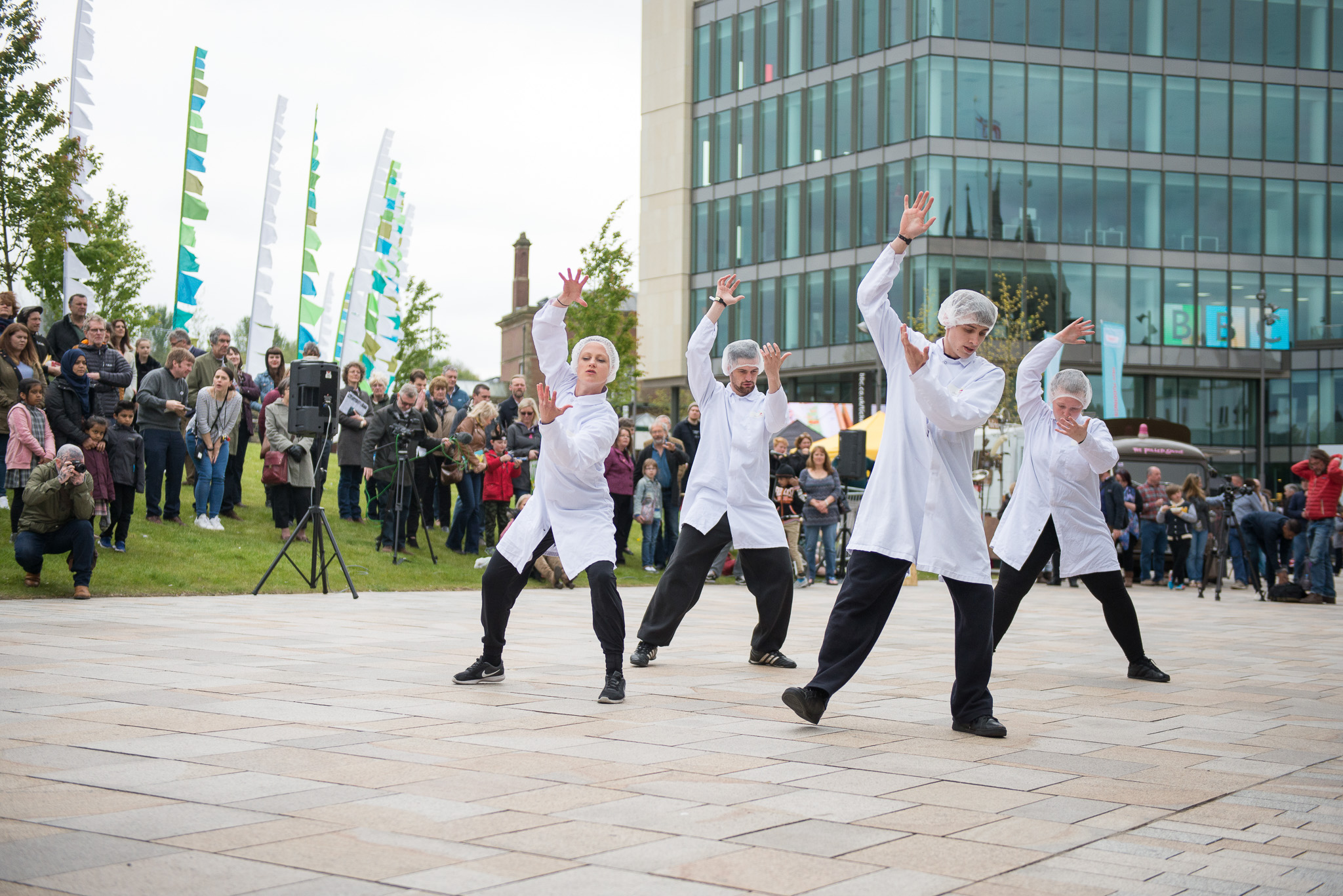 Four dancers wearing white lab coats and hairnets perform energetically in an outdoor plaza, while a crowd of onlookers watches in front of a modern glass building.