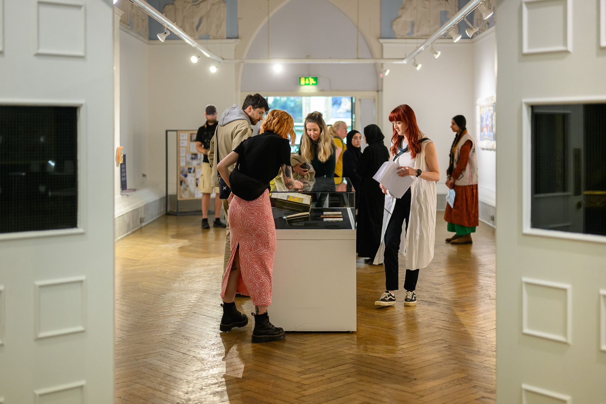 A group of people in a well-lit art gallery observe and discuss objects displayed in a glass case at the centre of the room. Some take notes, while others examine the exhibits closely.