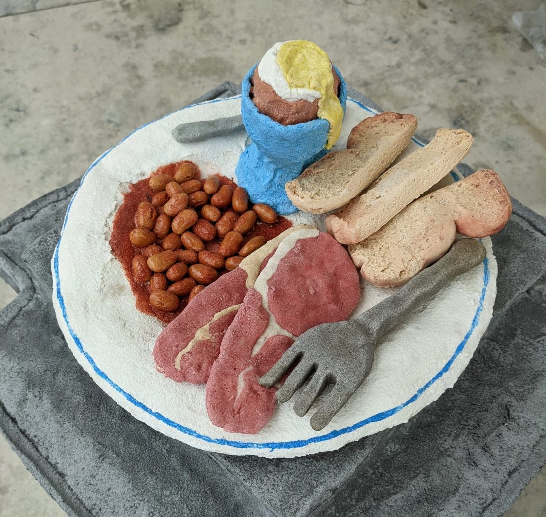 A textured, handmade plate of art resembling food, with clay-like beans, pinkish meat slices, bread, a boiled egg in a blue cup, and grey fork and knife, all placed on a grey surface.