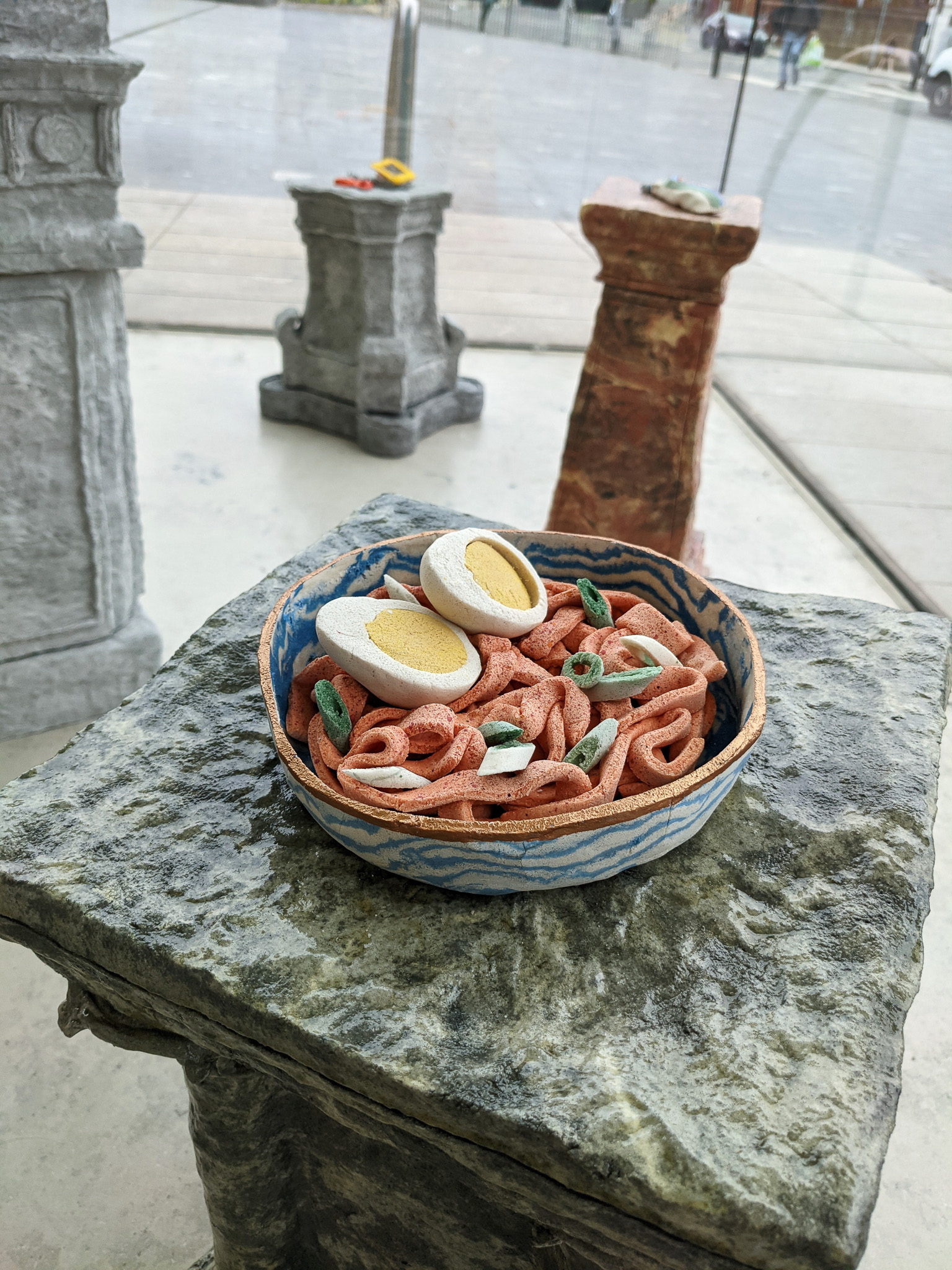 A ceramic bowl of sculpted noodles with boiled egg halves sits on a stone plinth, with other plinths holding small objects visible in the background near a window.