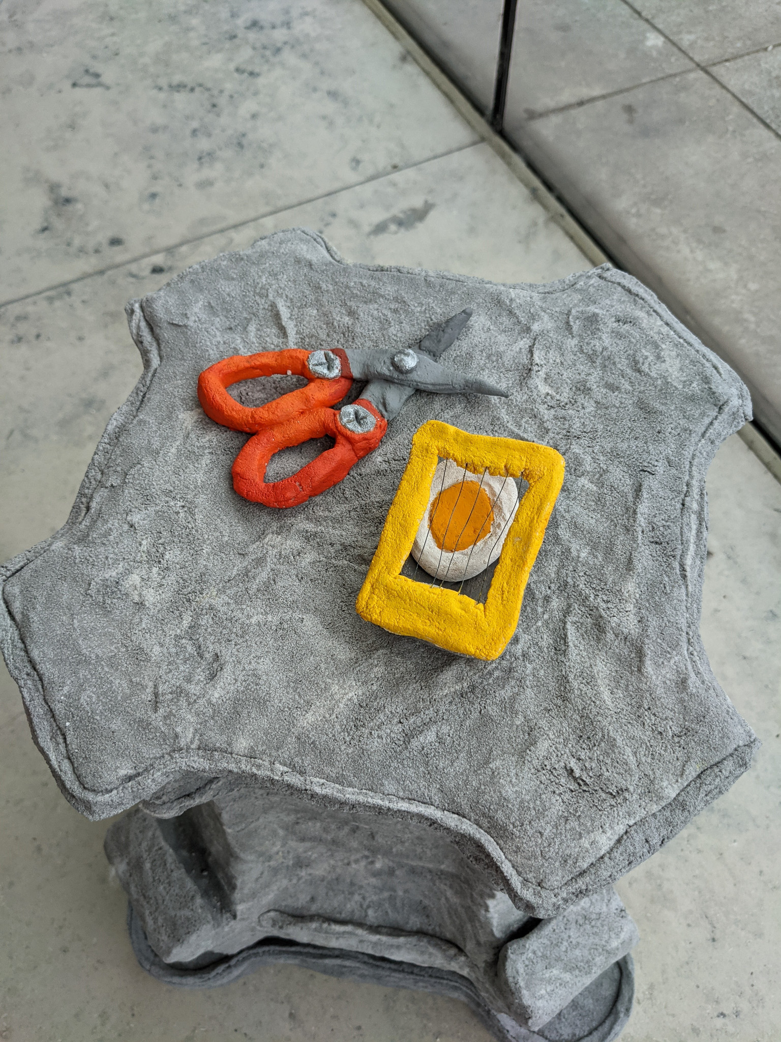 Stone-textured sculpture of orange scissors and a yellow egg slicer on a table.