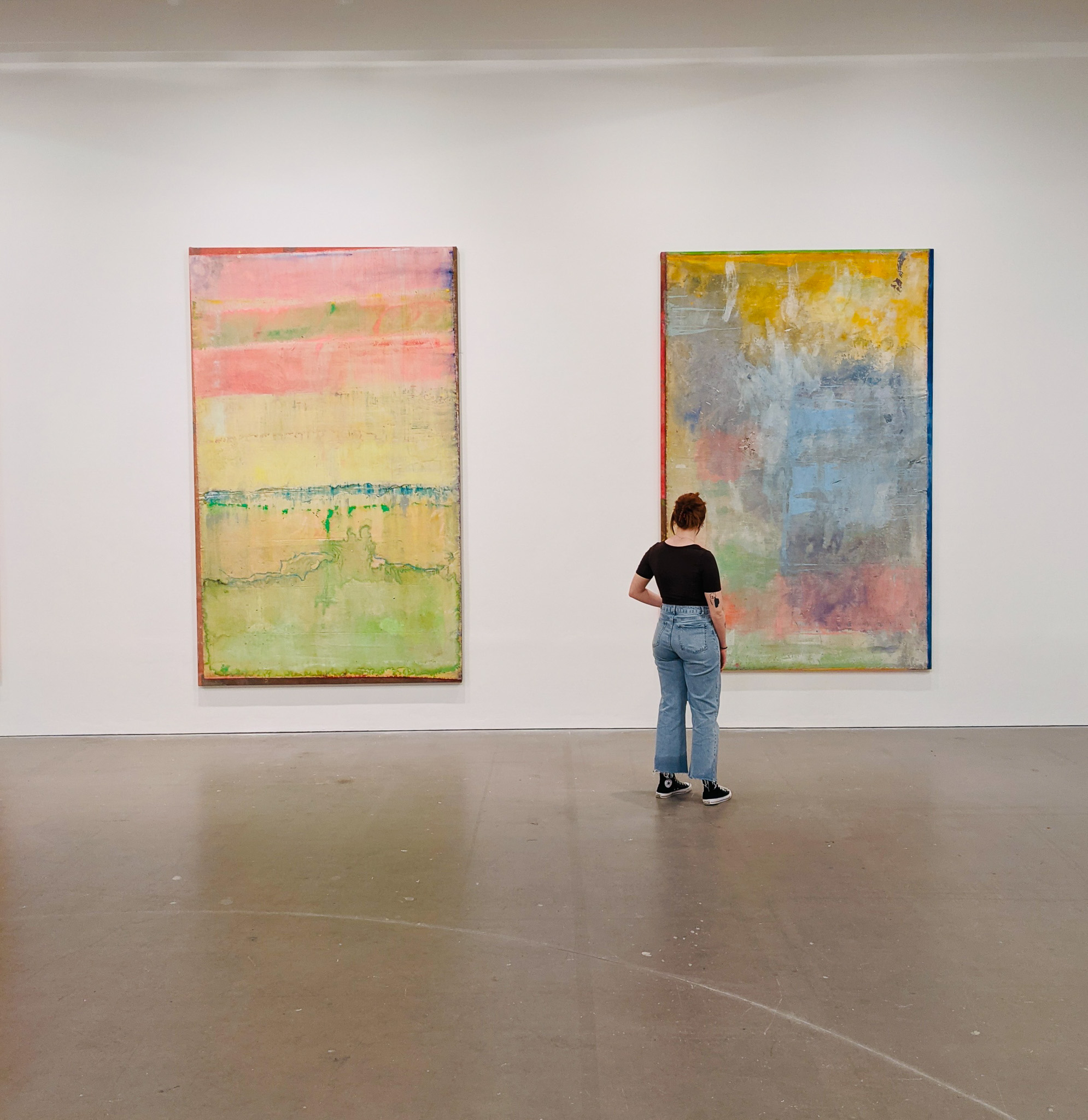 A person wearing a black shirt and blue jeans stands in an art gallery, looking at two large abstract paintings with pastel colours hanging on a white wall.