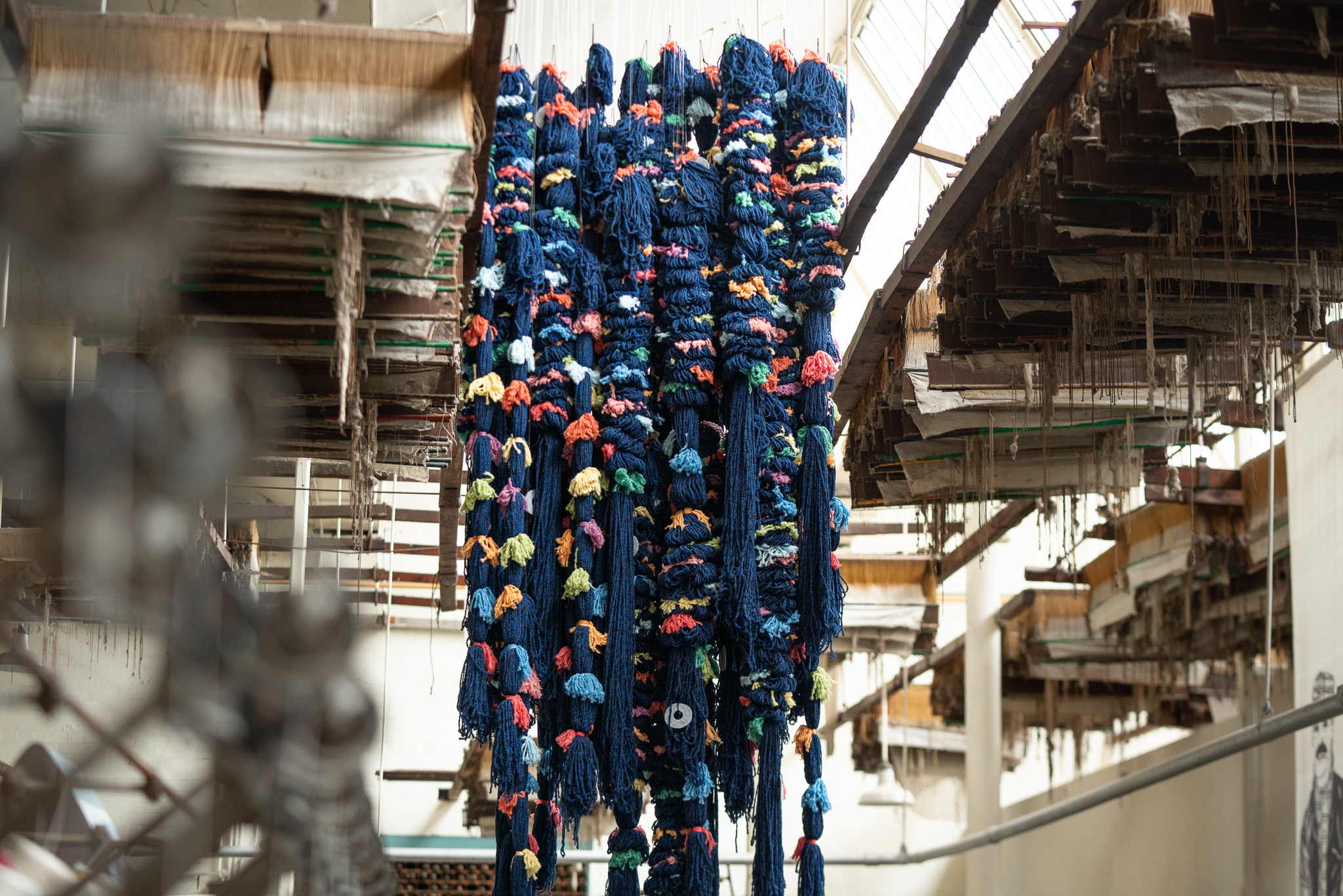 Bundles of blue yarn with colourful threads hanging from the ceiling in an industrial textile factory, surrounded by old wooden beams and machinery.