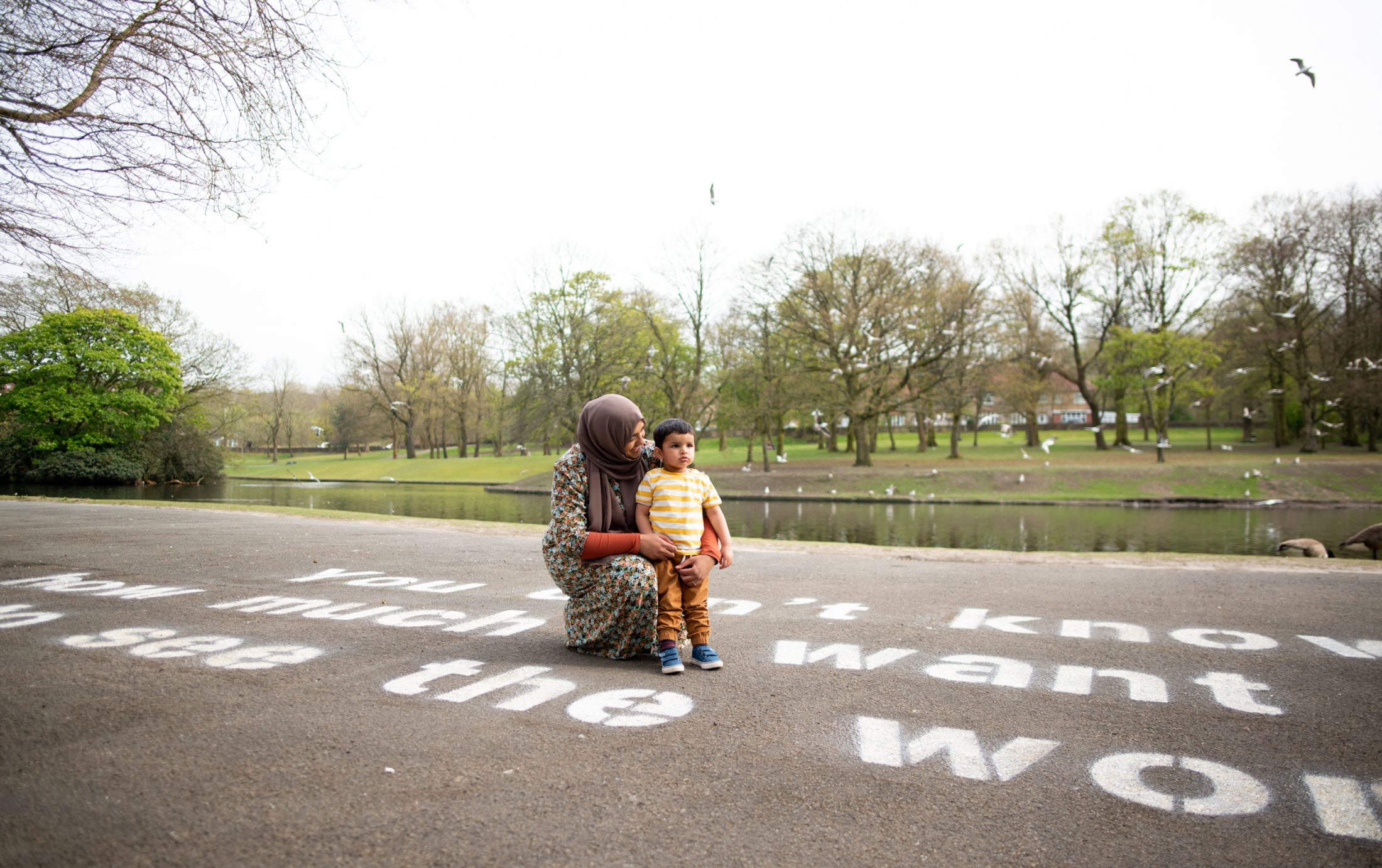 A woman wearing a patterned dress and headscarf kneels beside a young child in a park near a pond, with large white text painted on the path around them and trees in the background.