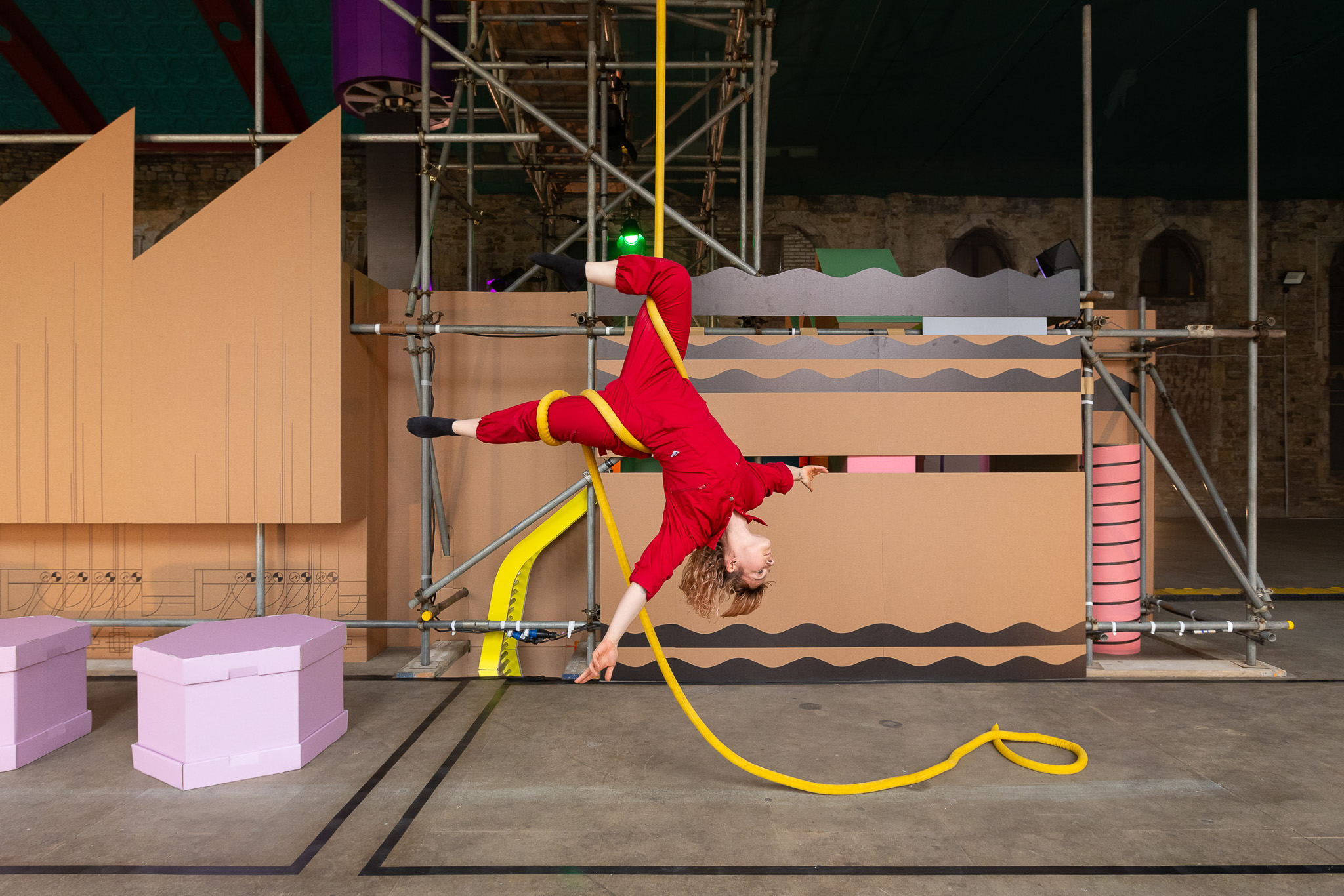 A person in a red jumpsuit performs an aerial acrobatic pose using a yellow rope, suspended upside down in a colourful industrial-like setting with cardboard structures and scaffolding.