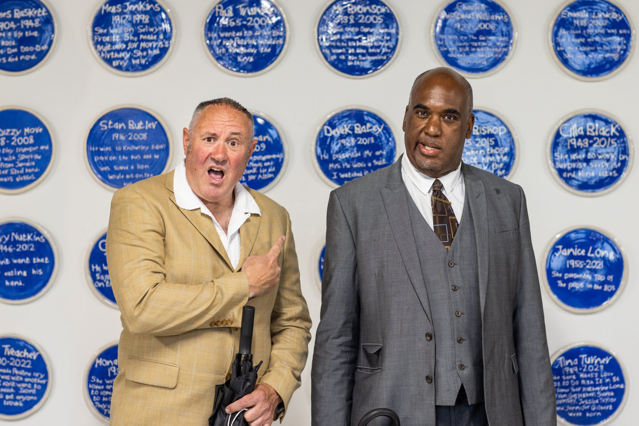 Two men in suits stand in front of a wall decorated with blue circular plaques featuring white text. One man gestures animatedly, while the other looks ahead with a neutral expression.