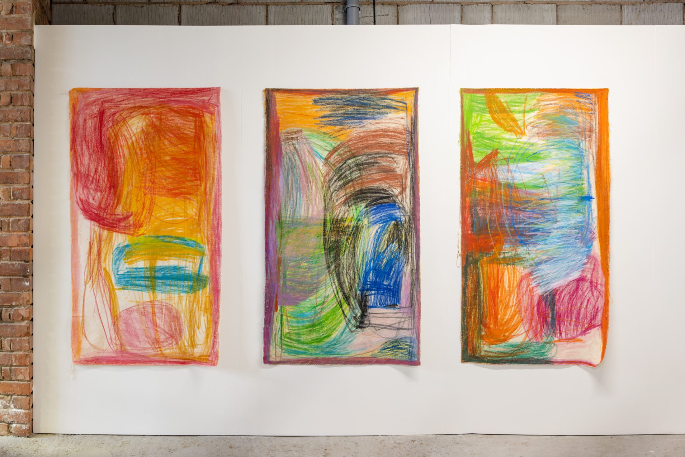 Three abstract, colourful paintings with expressive, layered strokes in red, orange, yellow, green, blue, and purple are displayed side by side on a white wall in an industrial-style space with a brick wall on the left.