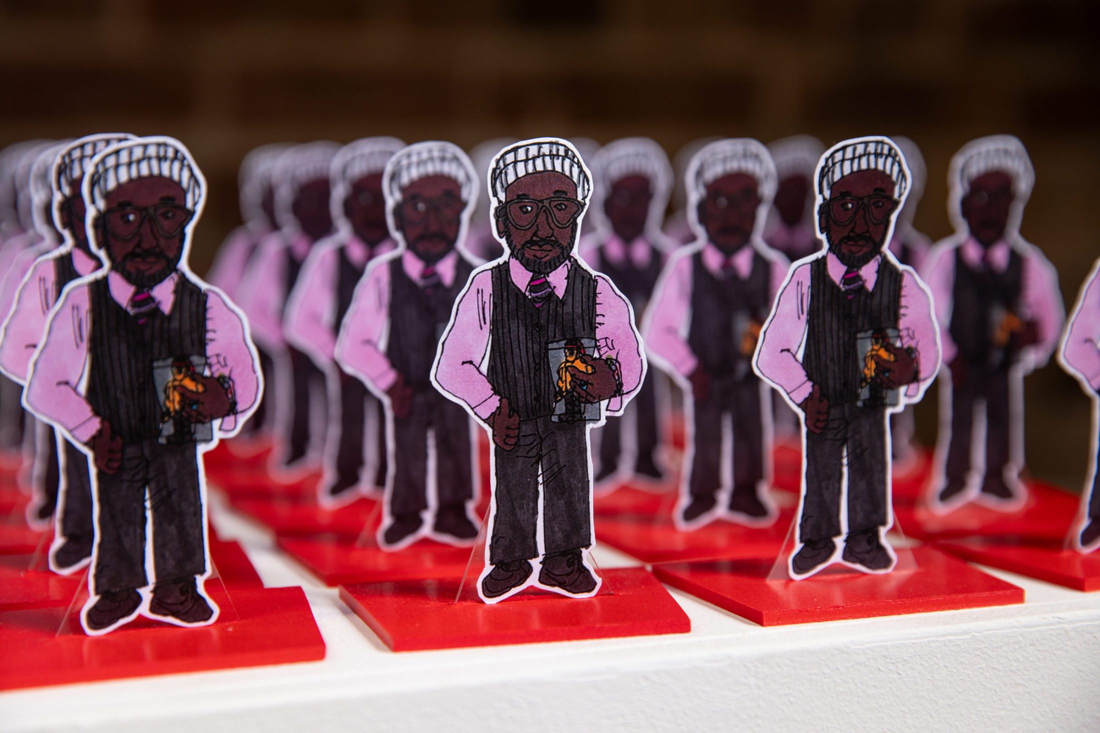 Multiple cut-out figures of a man wearing glasses, a white hat, pink shirt, and black waistcoat, holding a drink, are arranged in rows on red bases against a blurred background.