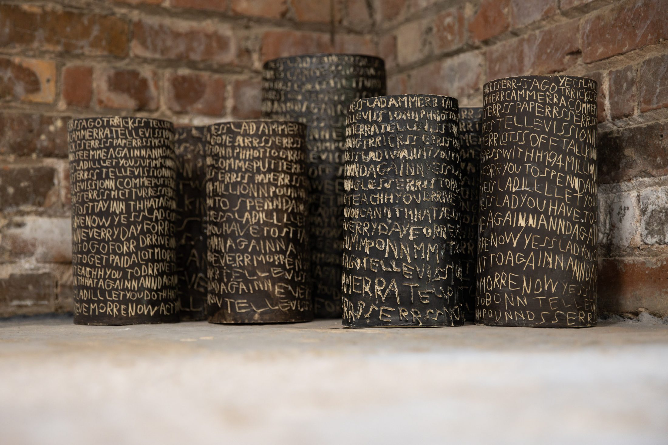 Five cylindrical sculptures with inscribed text stand against a brick wall.