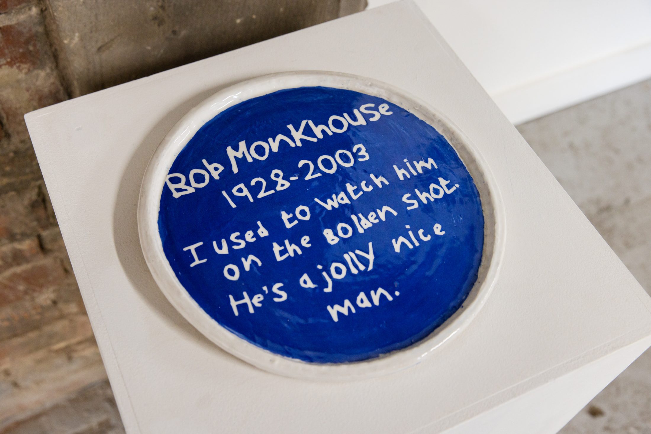 Blue ceramic plate on a white pedestal, inscribed with handwritten text in white, expressing admiration for Bob Monkhouse.