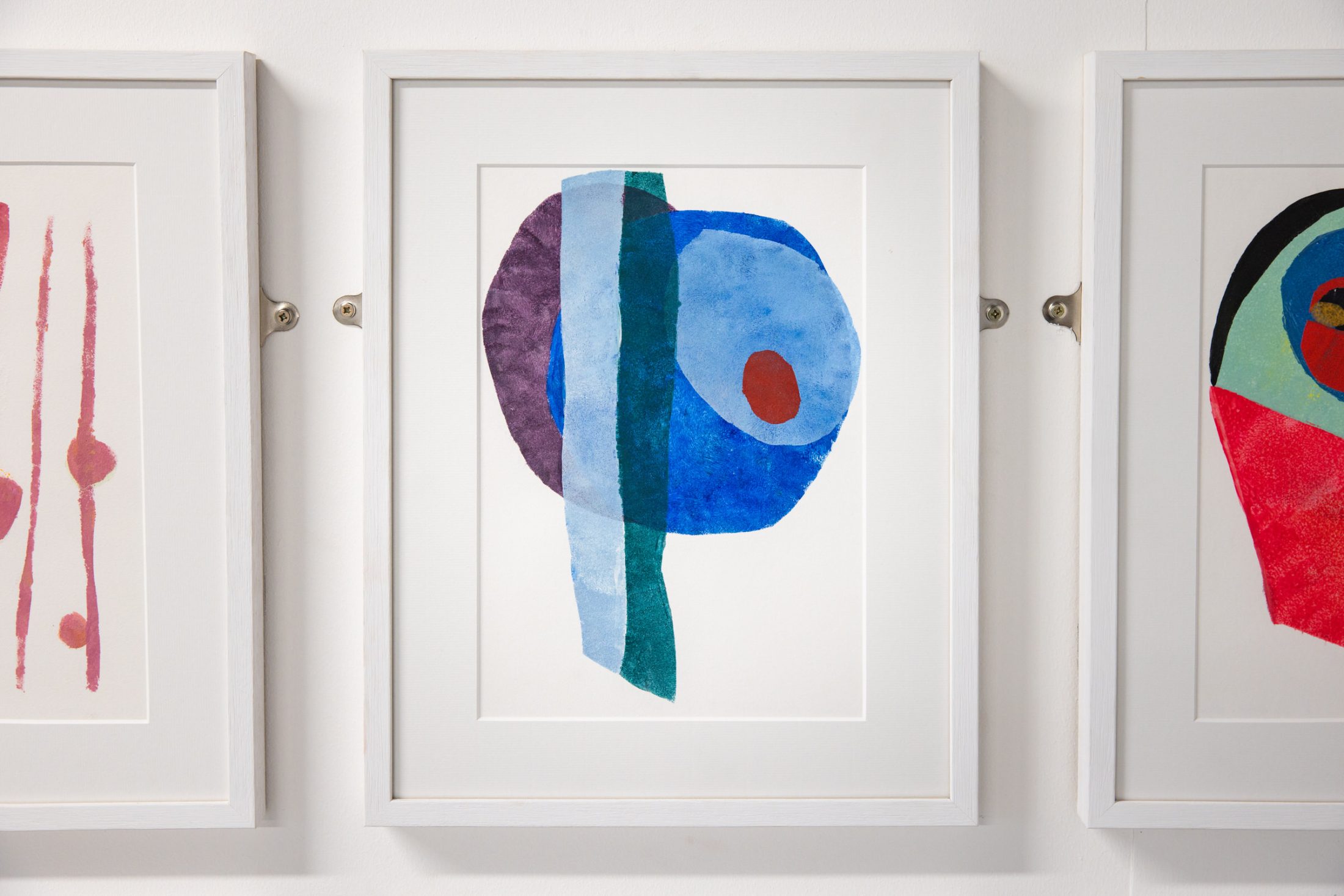 Abstract painting in a white frame features blue, teal, and maroon shapes overlapping. The artwork is part of a series on a gallery wall.