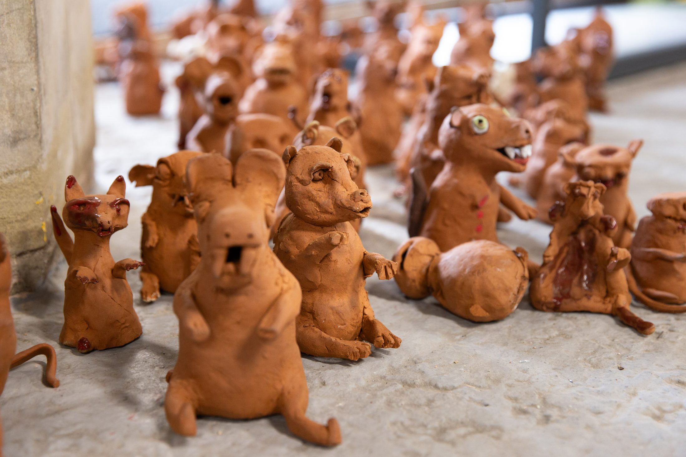 A group of small, handmade clay animal sculptures with rough, cartoonish features are arranged on a concrete floor. The figures vary in size and expression. Some are upright, while others are sitting or lying down.