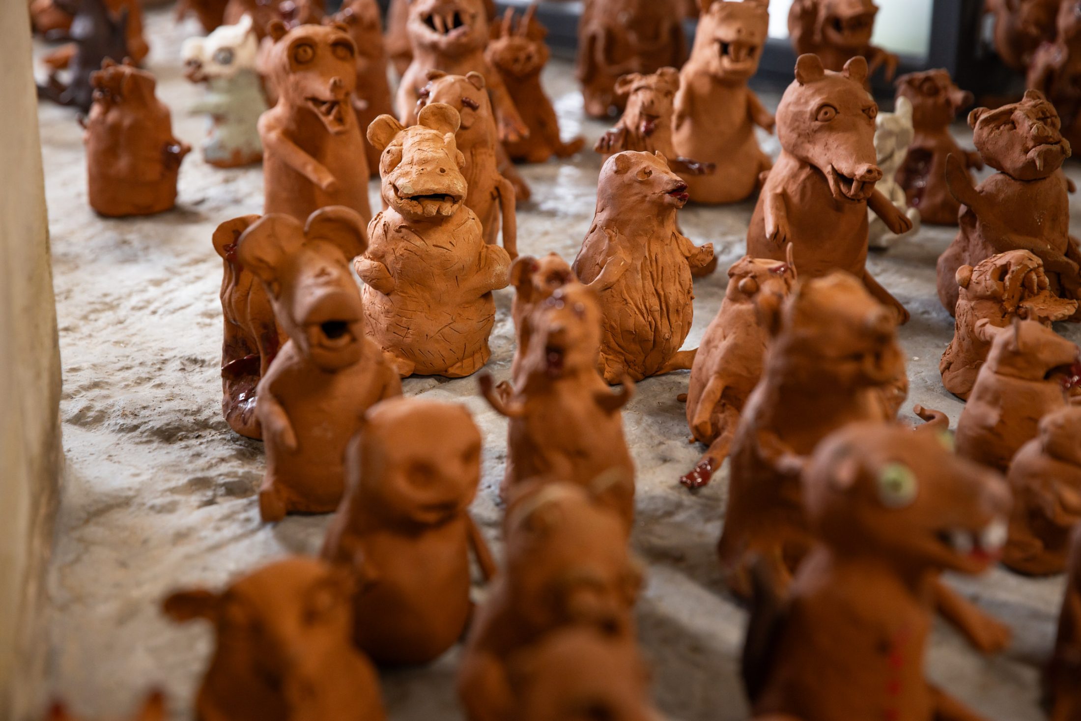 Several handmade clay animal figurines with exaggerated expressions are arranged on a sandy surface, closely grouped together and facing different directions.