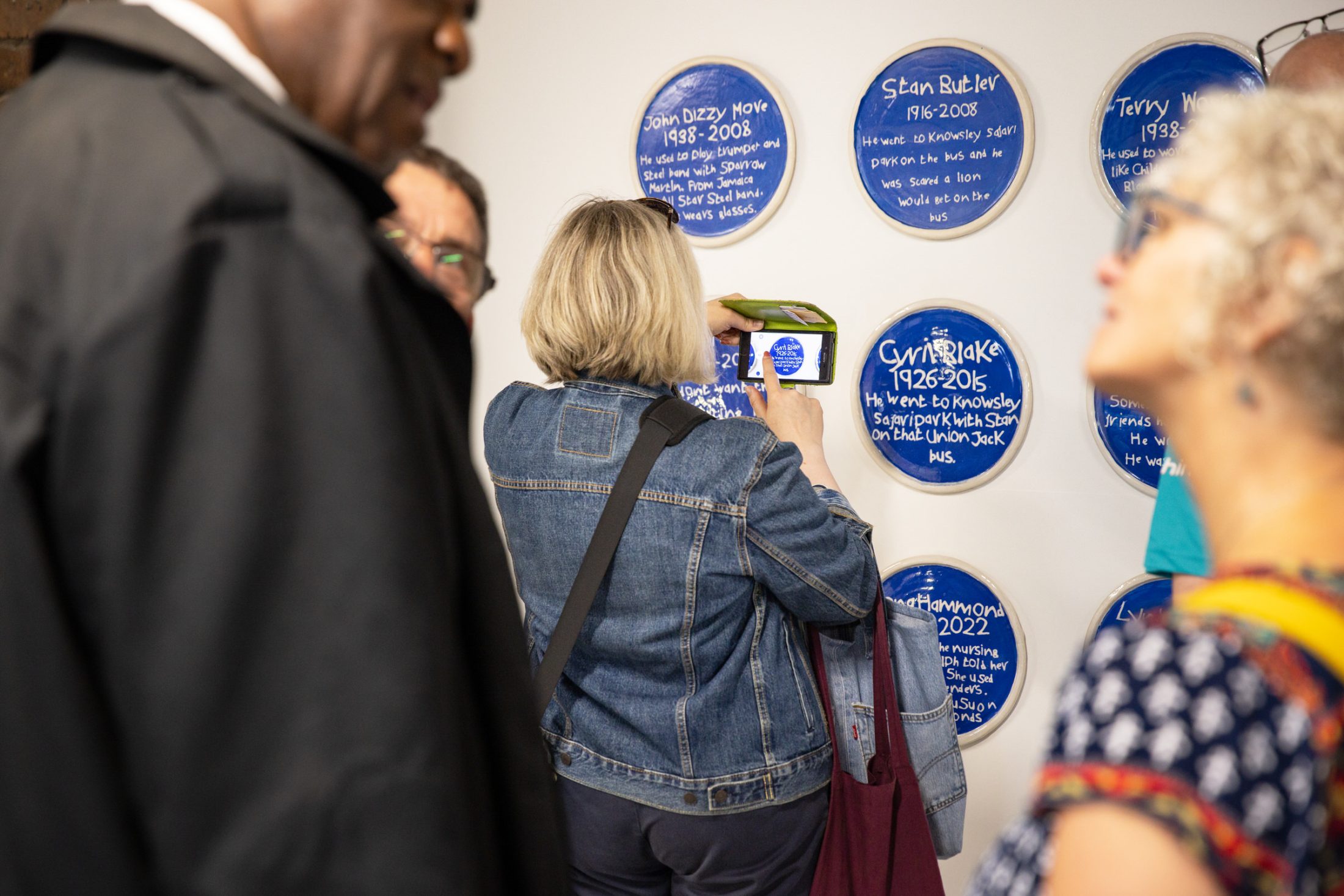 People in a gallery observe blue circular plaques with white text. A woman in a denim jacket photographs a plaque.