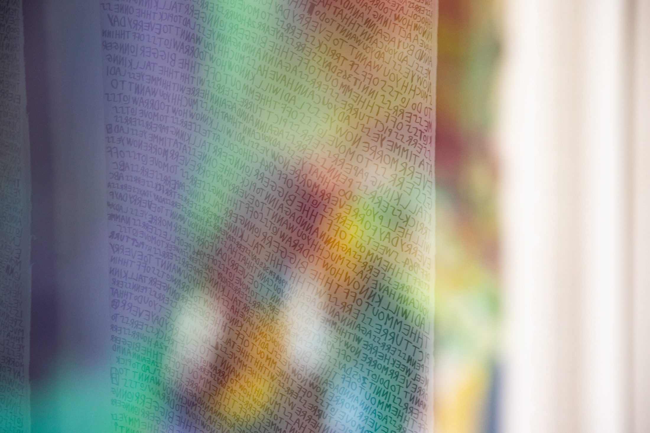 A close-up of a translucent sheet covered in handwritten text, with colourful, blurred shapes visible behind the sheet. The writing appears reversed and is densely packed across the surface.