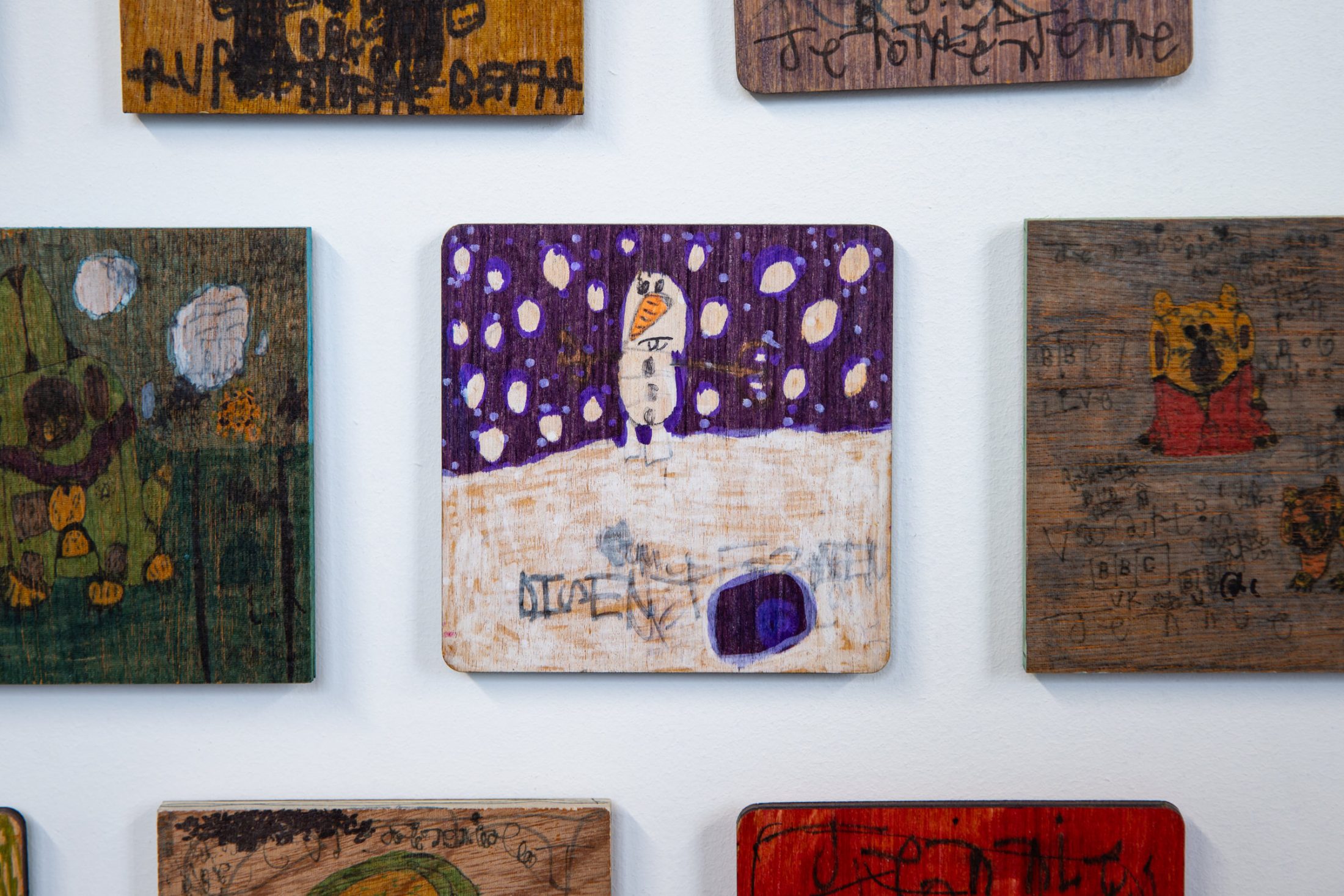 Artwork on wood features a snowman with an orange nose under a purple, snow-filled sky. Other pieces surround it on a white wall.