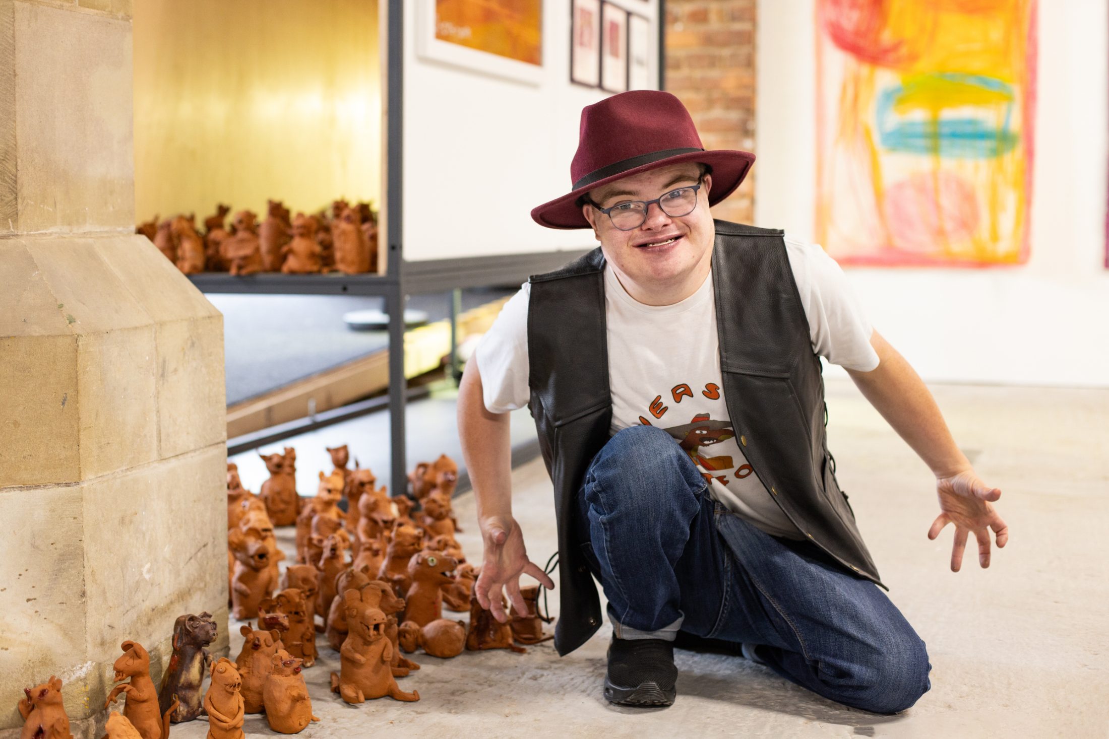 A person in a burgundy hat and leather vest crouches joyfully beside a collection of clay weasels sculptures in a bright art gallery setting.