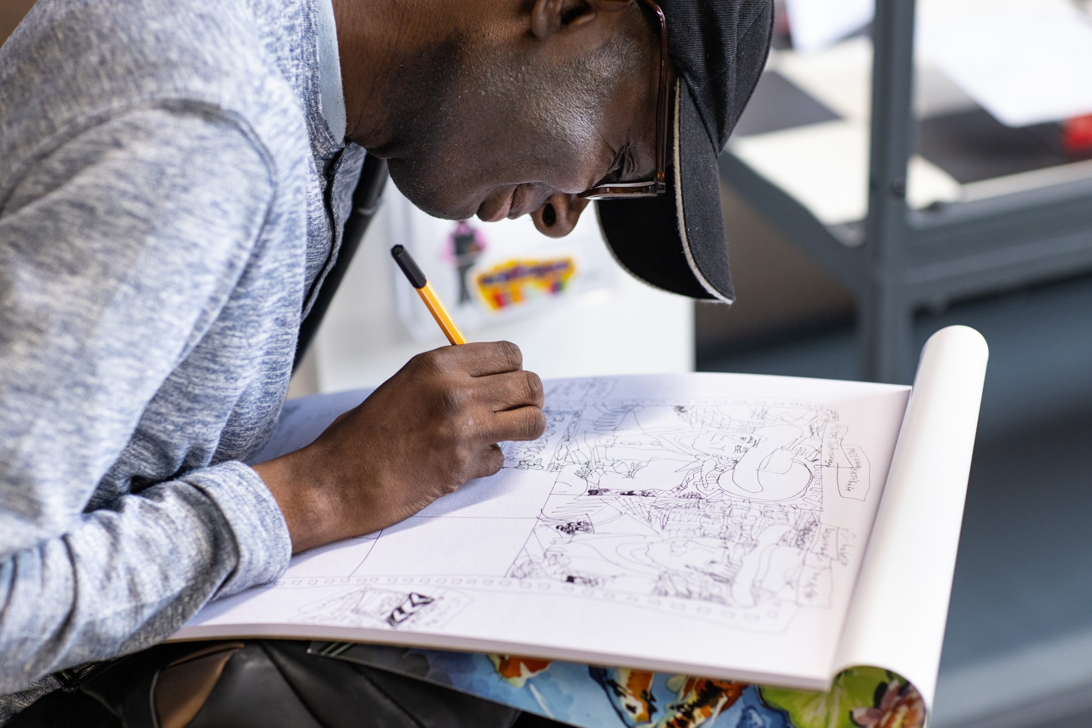 A person in a cap and glasses intensely focuses on drawing with a pencil on a large sketchpad filled with intricate comic illustrations.