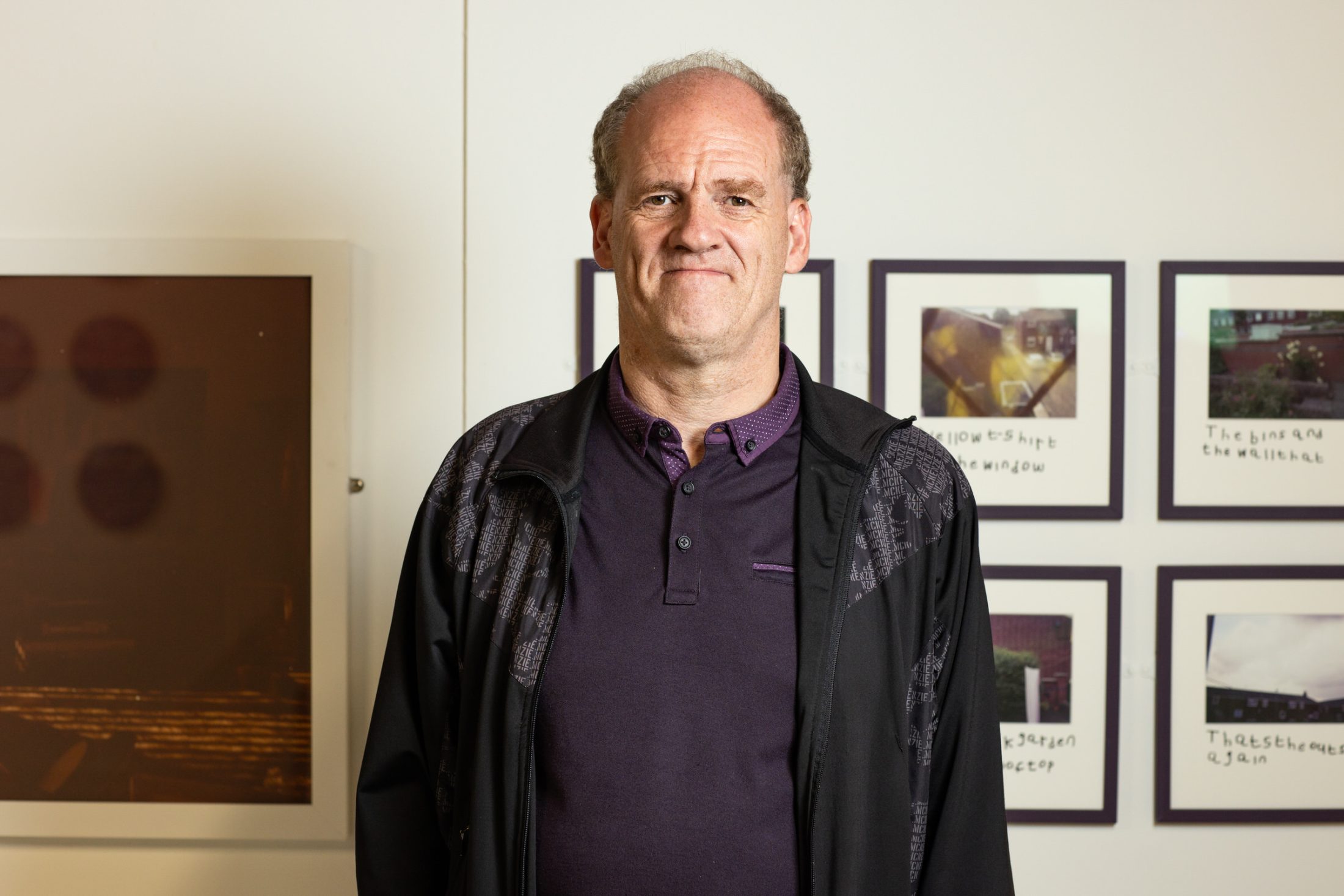 A man in a black jacket and purple shirt stands in a gallery space with framed photographs on the wall behind him.