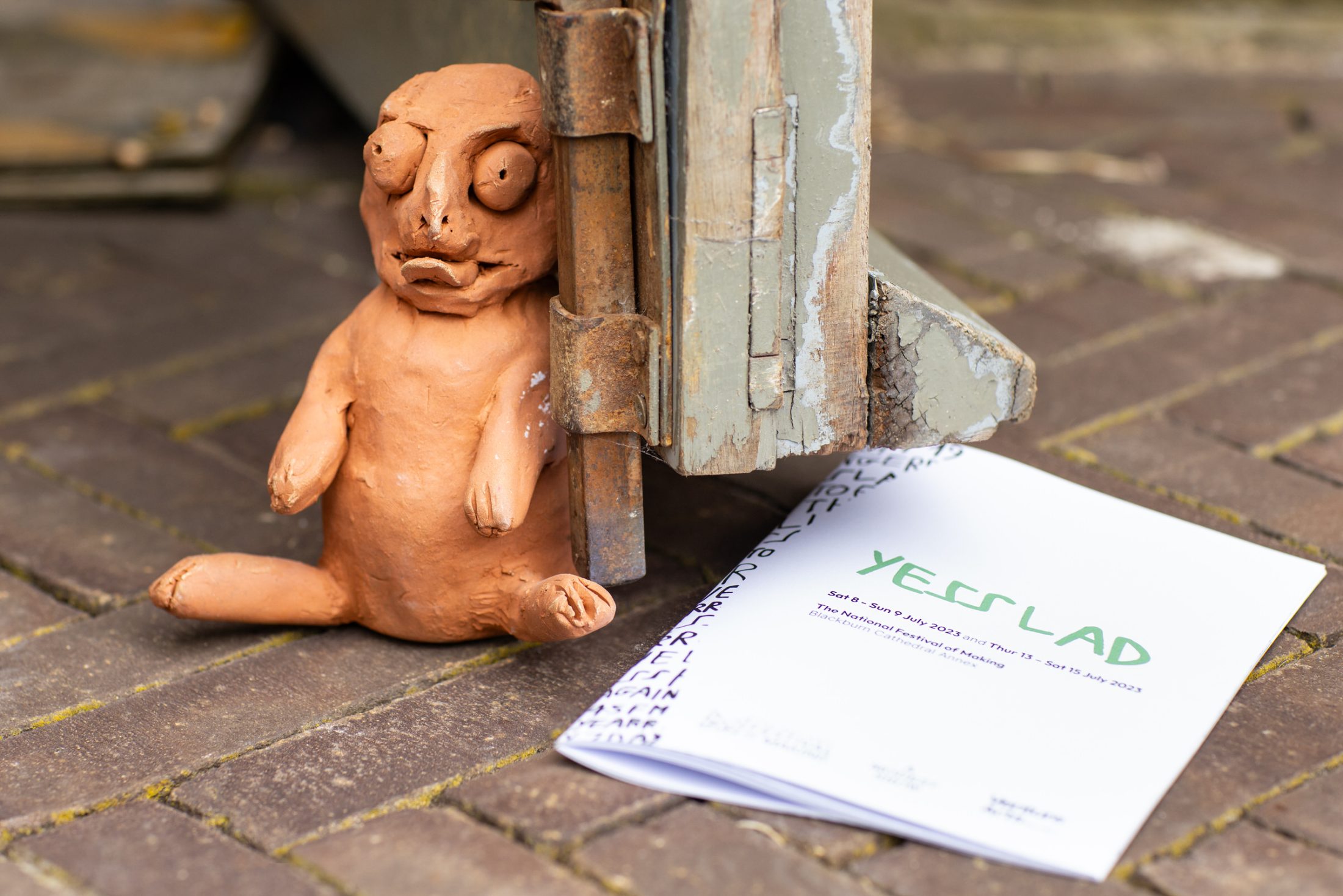 A small, roughly sculpted clay figure sits on the ground next to a weathered wooden door. A white booklet titled YESSLAD lies beside the figure on a brick pavement.