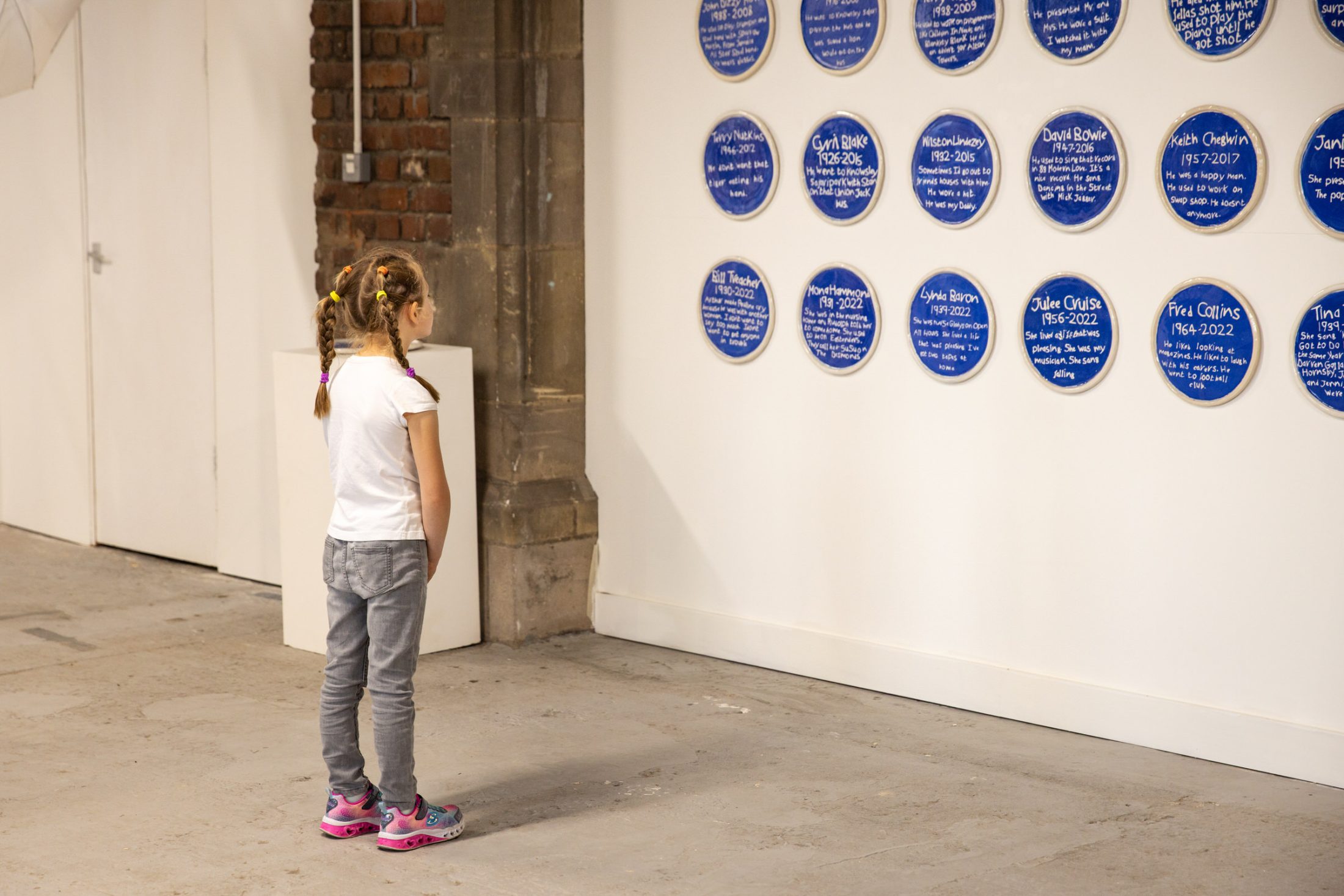 A young girl with braided hair stands in a gallery, looking at a wall displaying blue circular plaques with white text.