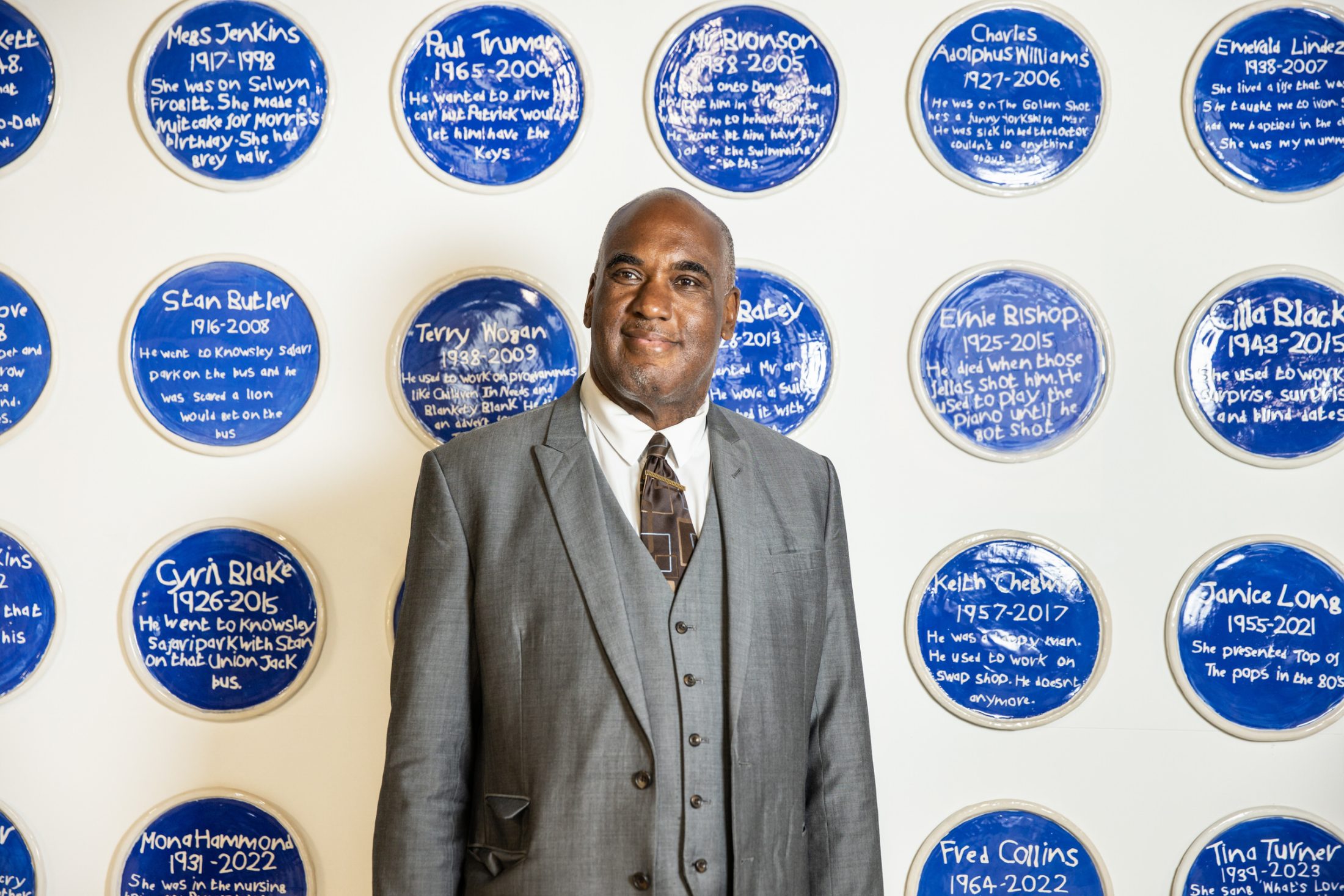 A man in a grey suit stands smiling against a wall decorated with blue circular plaques featuring white handwritten text.