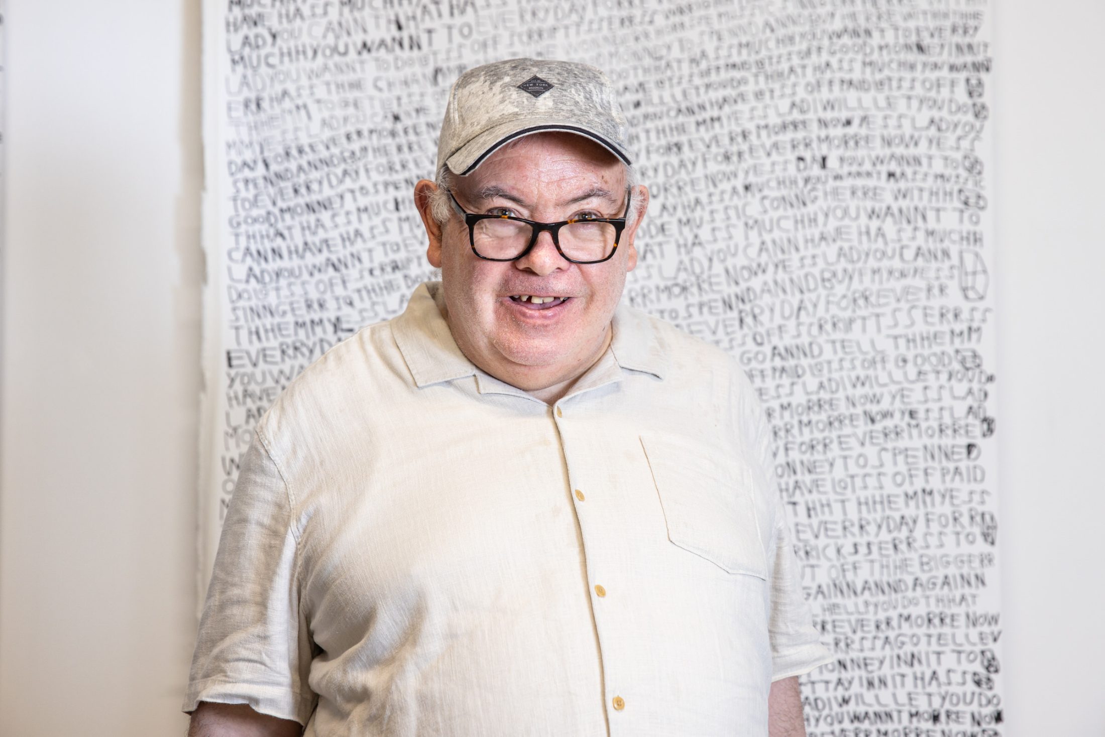 A man wearing glasses, a light beige shirt, and a cap stands smiling in front of a large artwork filled with handwritten text.