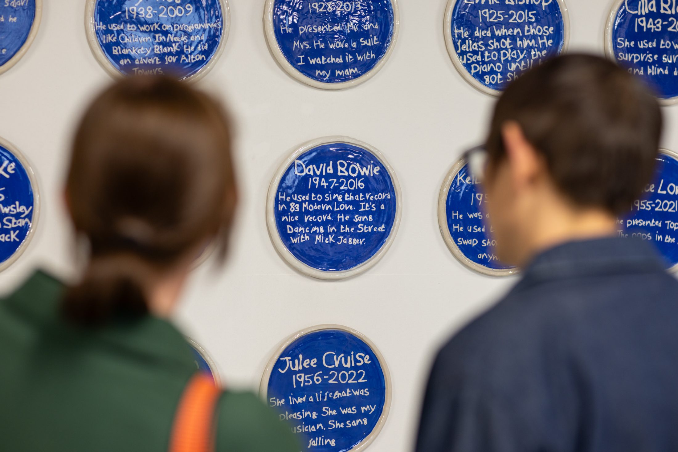 Two people view a wall display of blue circular plaques with white writing, each commemorating different individuals with their names, dates, and brief descriptions.