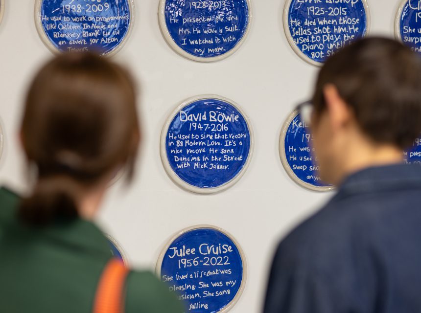 Two people view a wall display of blue circular plaques with white writing, each commemorating different individuals with their names, dates, and brief descriptions.