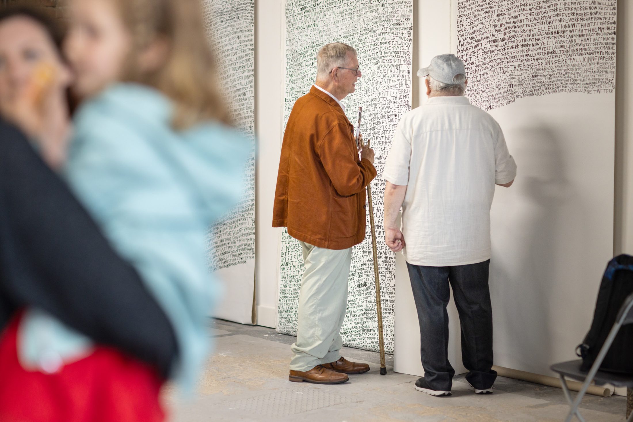 Two older men stand and talk in front of large white panels covered in handwritten text. One man holds a walking stick. The scene is indoors, with a blurred woman and child in the foreground on the left.