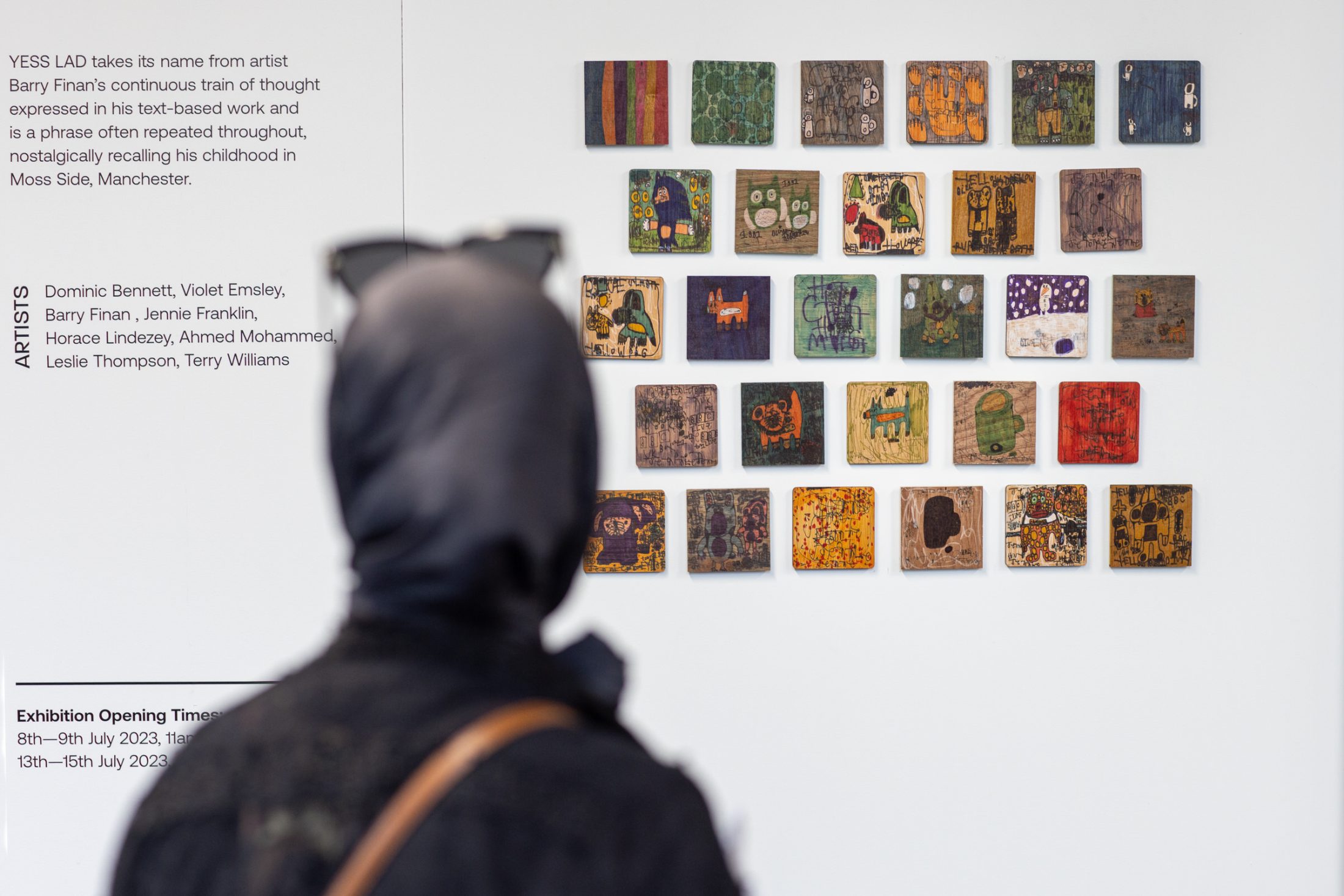 A person in a black hooded jacket observes a gallery wall with a grid of colourful abstract art pieces. Texts about the exhibition are visible on the wall.