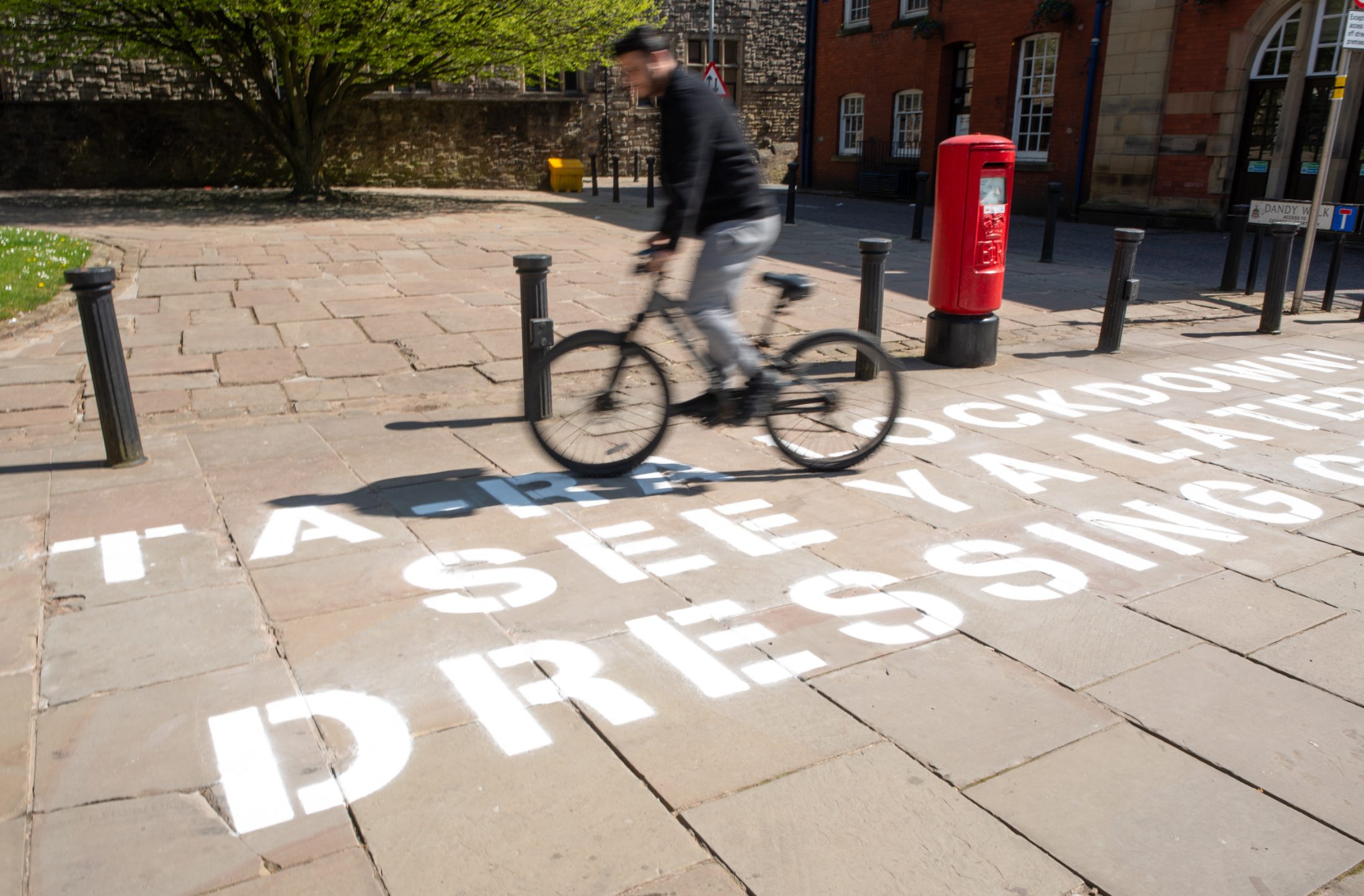 A cyclist rides past large white text painted on a sunny cobblestone path, reading 