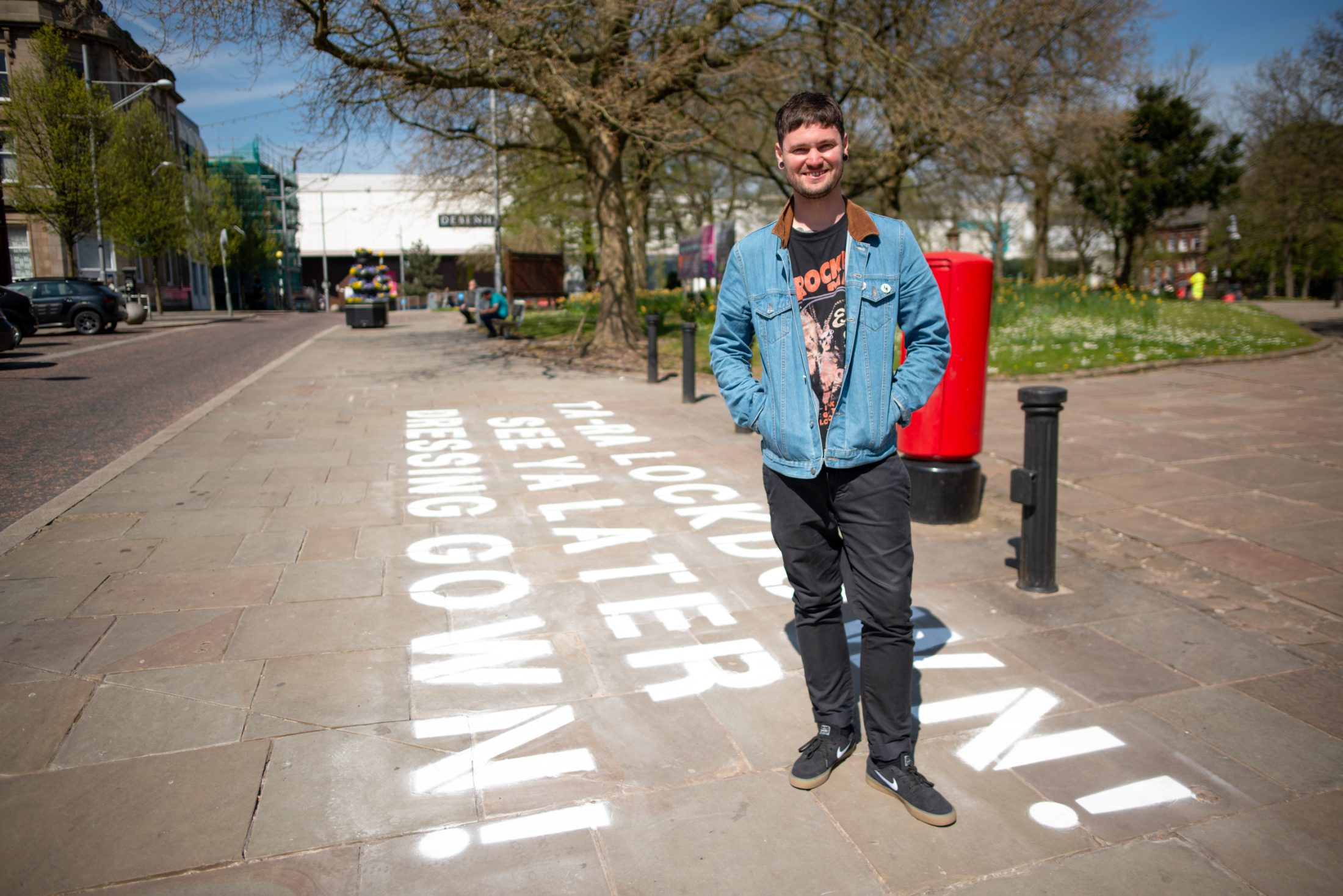 A young man in a denim jacket and graphic T-shirt stands smiling on a sunny street, next to a red postbox. Large white letters painted on the pavement read, I am lockdown! See you later, dressing gown!.