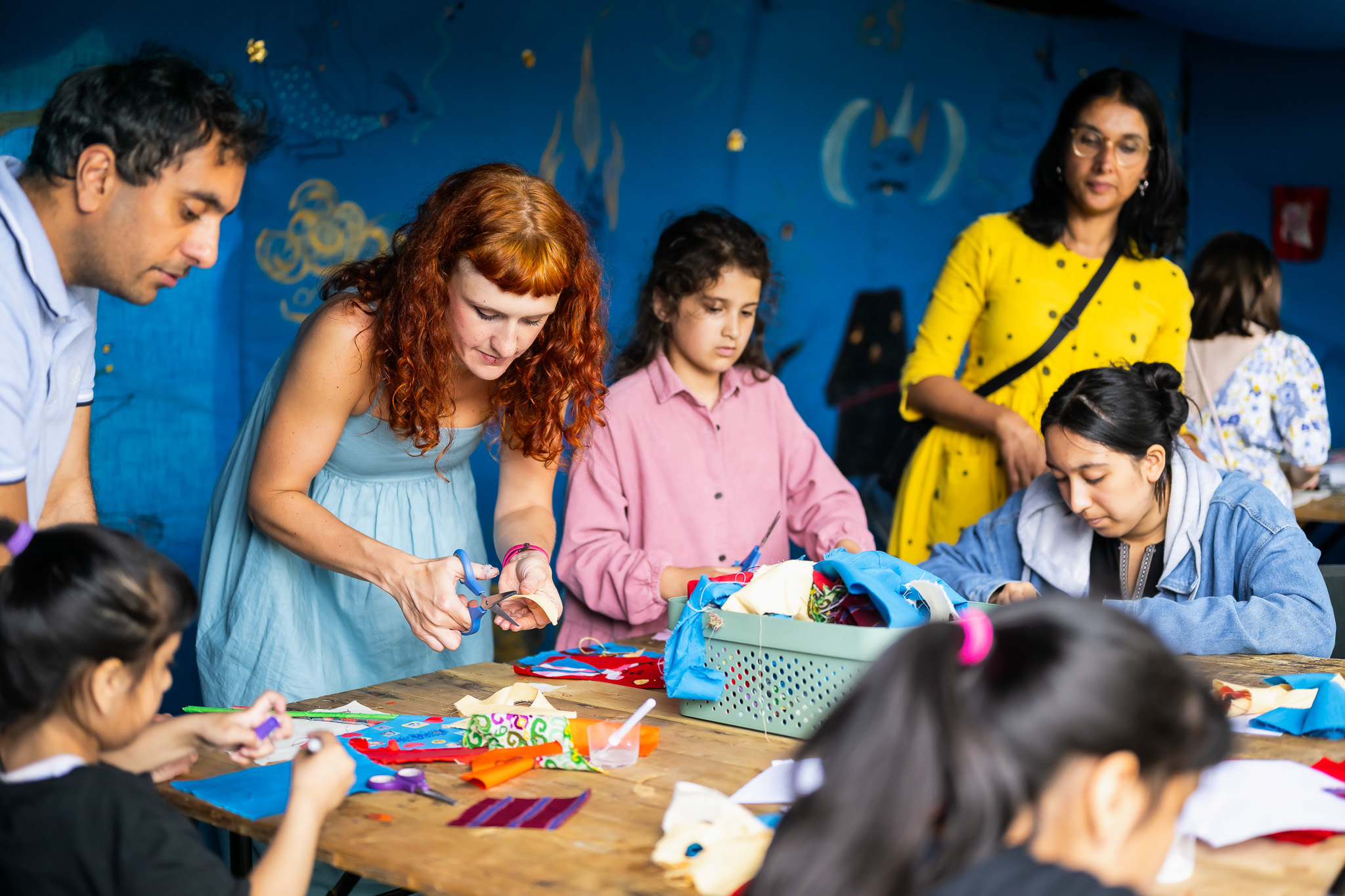 A group of adults and children sit and stand around a table, engaged in arts and crafts activities, cutting and assembling colourful materials in a bright, cheerful indoor setting.