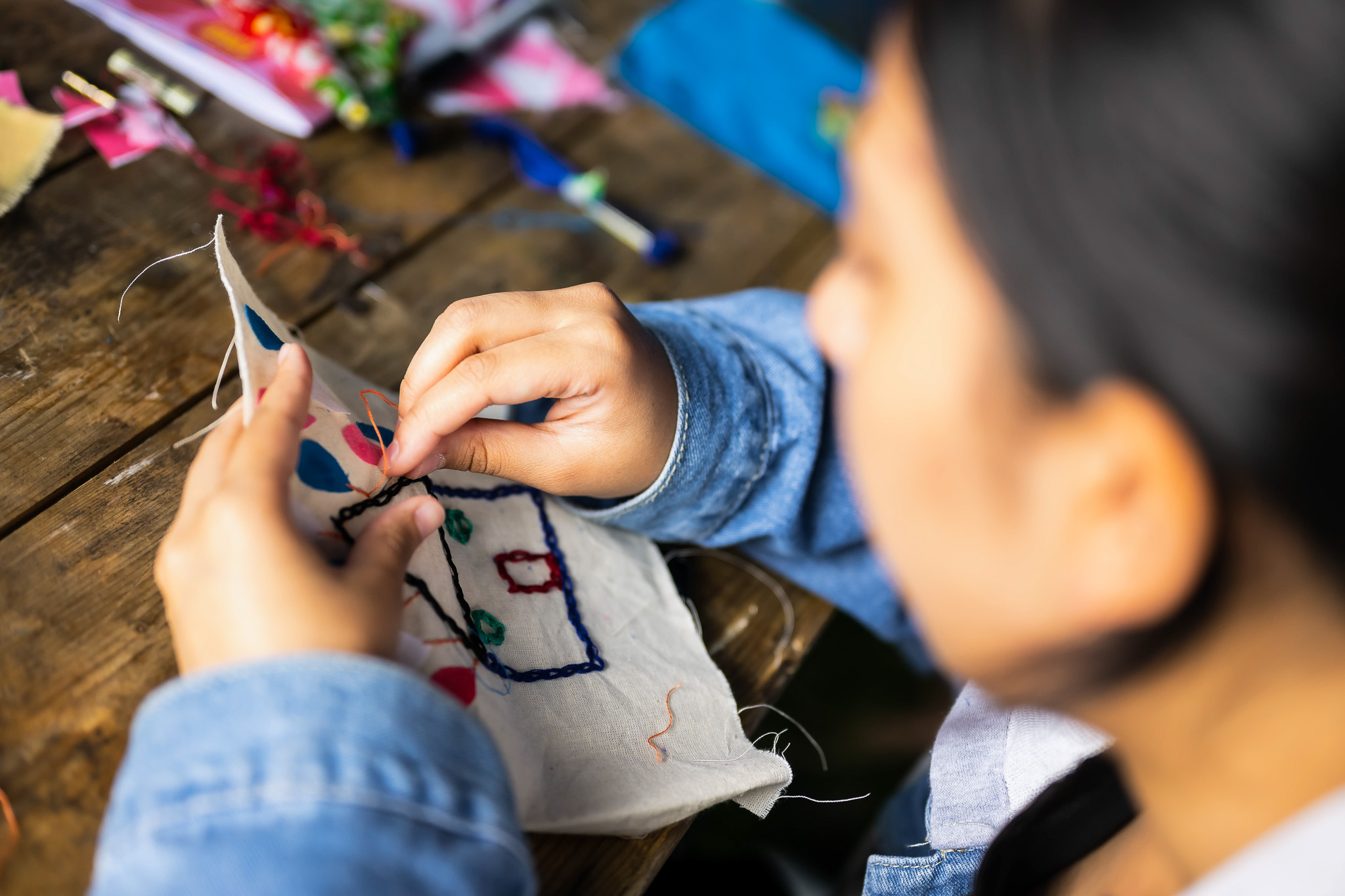A person wearing a blue denim jacket carefully hand-stitches colourful thread patterns onto a piece of fabric, working at a wooden table with embroidery materials scattered nearby.