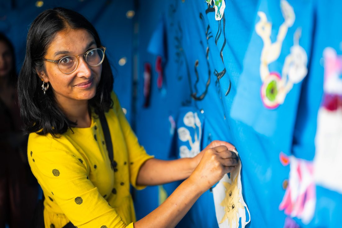 A woman in glasses and a yellow shirt pins paper cut-outs onto a blue display board, looking at the camera. Bright artwork and decorations are visible in the background.