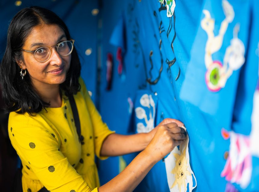 A woman in glasses and a yellow shirt pins paper cut-outs onto a blue display board, looking at the camera. Bright artwork and decorations are visible in the background.