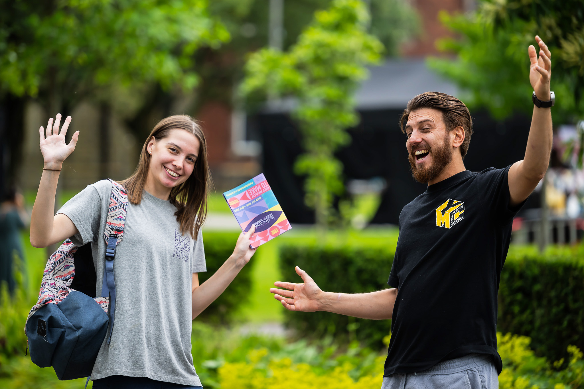 Two young adults stand outdoors, smiling and waving. The woman holds colourful leaflets and carries a rucksack, while the man gestures with open arms. Green trees and bushes are visible in the background.