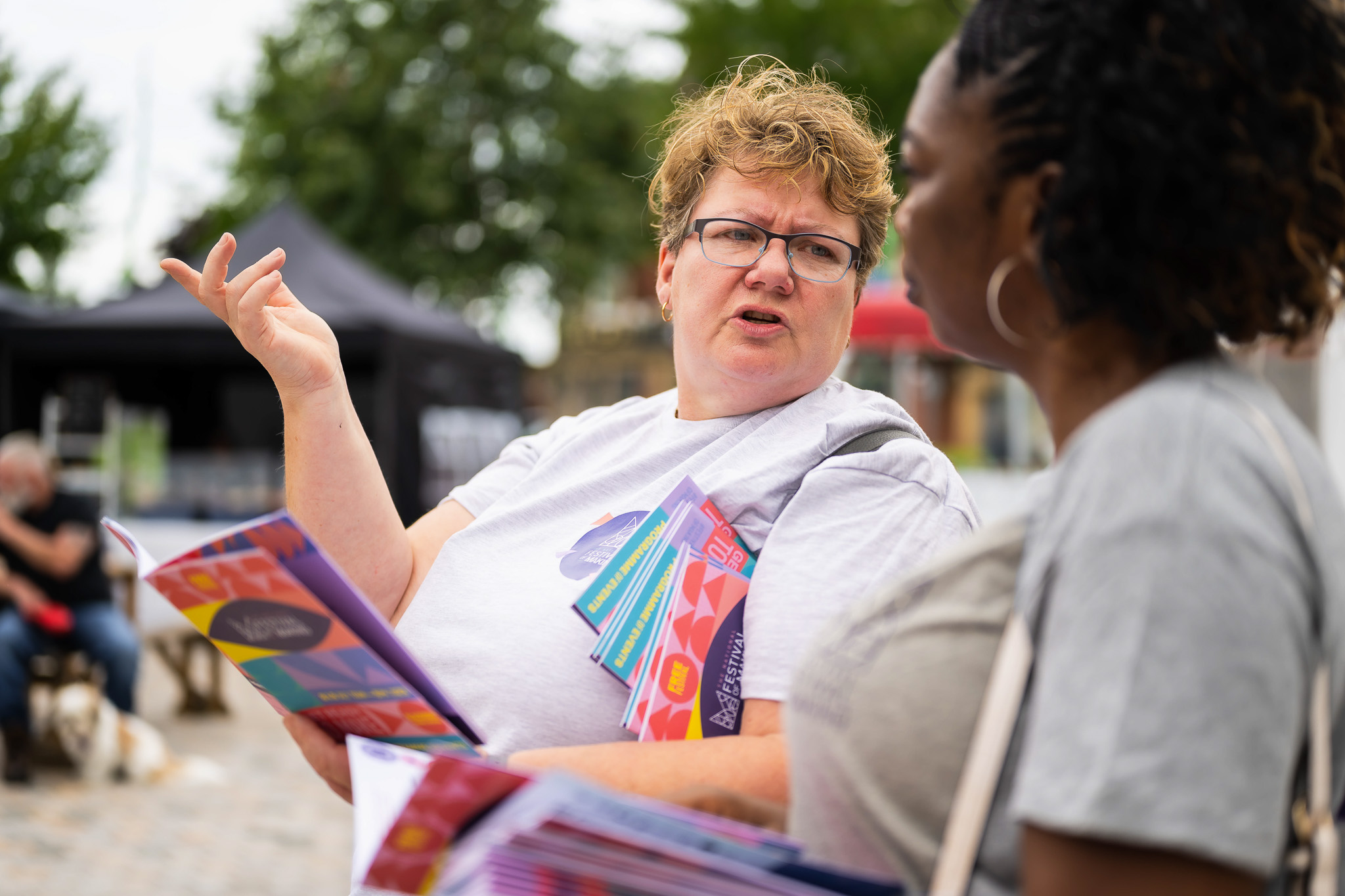 A woman in a white shirt gestures whilst talking to another woman outdoors, holding colourful pamphlets. They appear to be at an event or market, with marquees and people in the background.