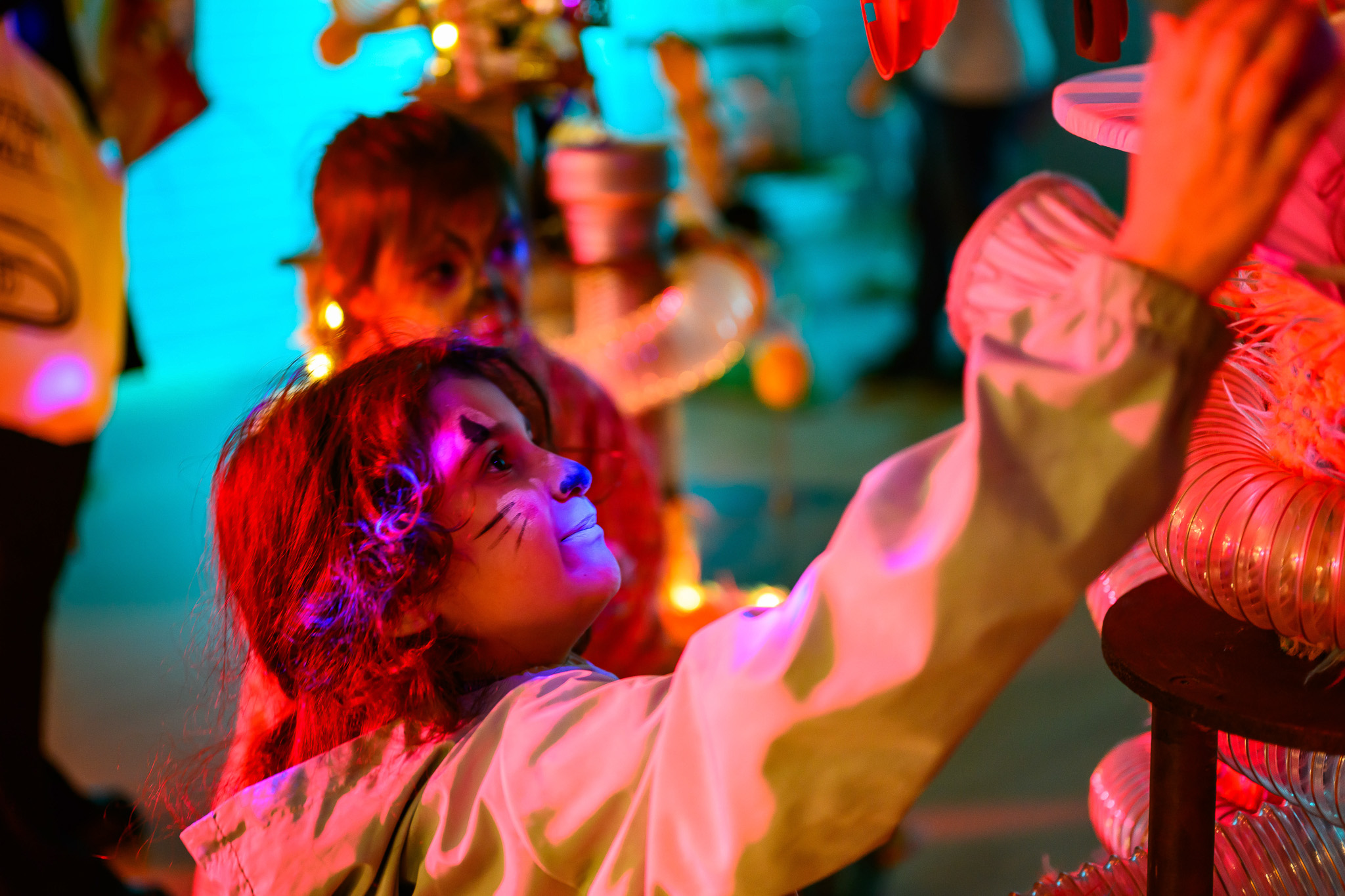 A child with face paint resembling a cat reaches towards a display, surrounded by colourful lights, while another child stands behind, both appearing to enjoy a festive event.