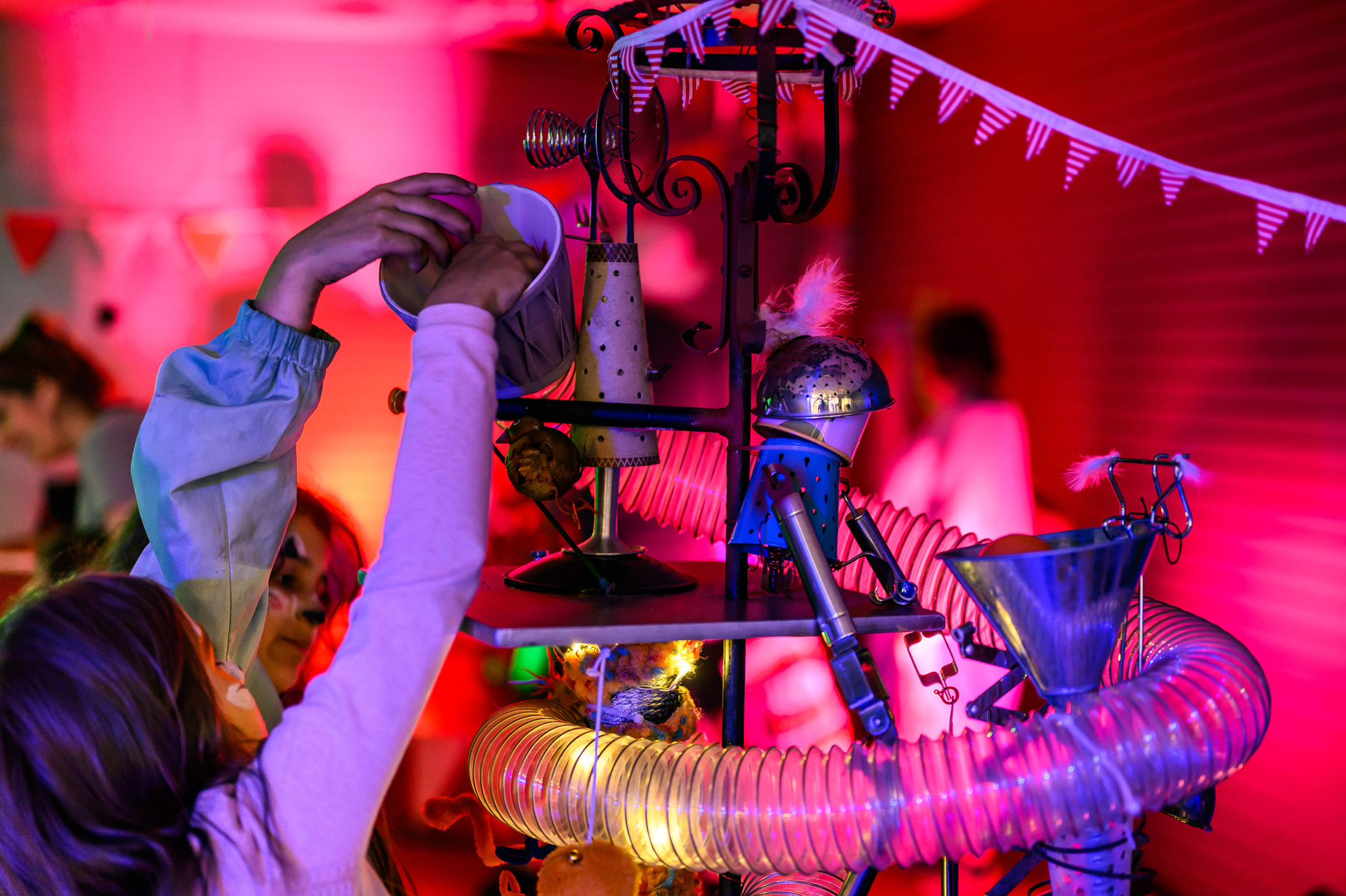 Two children interact with a colourful, whimsical contraption made of tubes, funnels, and decorations in a brightly lit room with pink and purple lighting. Bunting hangs in the background, creating a festive atmosphere.