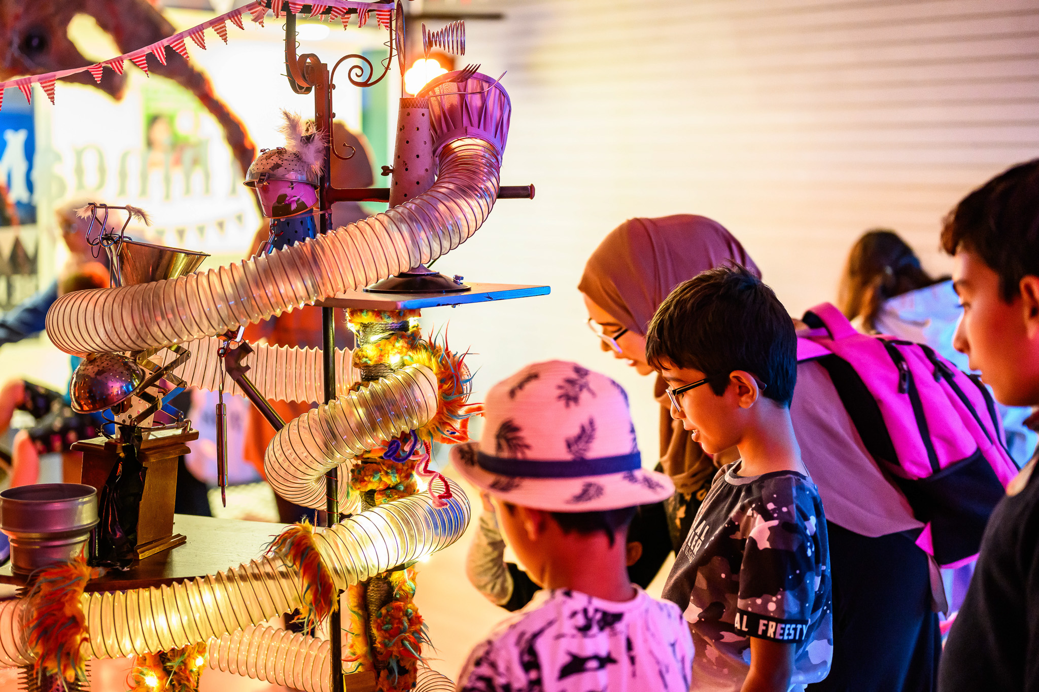 Children and a woman look at a colourful, whimsical machine with tubes and lights at an indoor event, surrounded by a festive atmosphere.