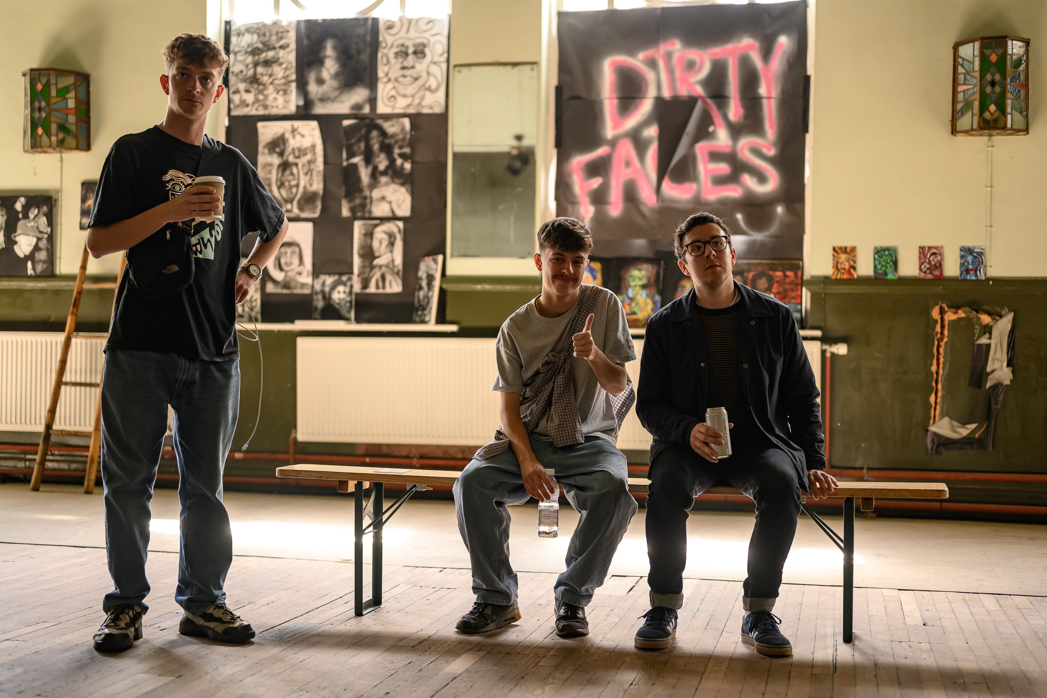 Three young men pose in an art room with drawings on the wall and a large DIRTY FACES sign. Two sit on a bench, one holding up his finger, whilst the third stands holding a drink. The room has wooden floors and bright lighting.